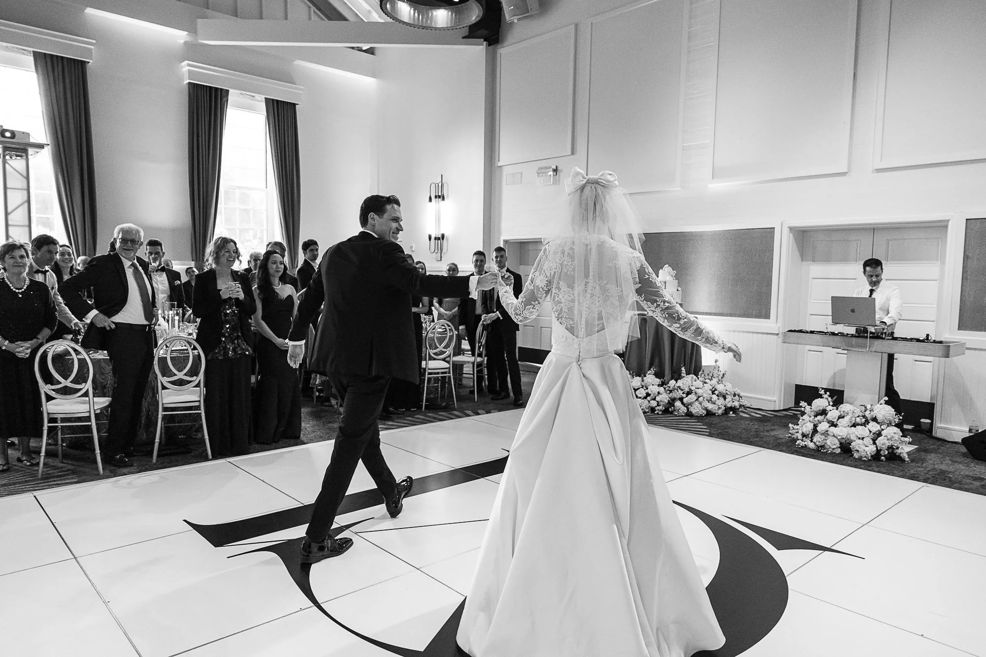 A wedding reception with a bride in a long white dress and veil dancing with a man in a tuxedo on a dance floor. Guests are watching and smiling, seated at tables with floral arrangements in a well-lit room with tall windows and dark curtains.