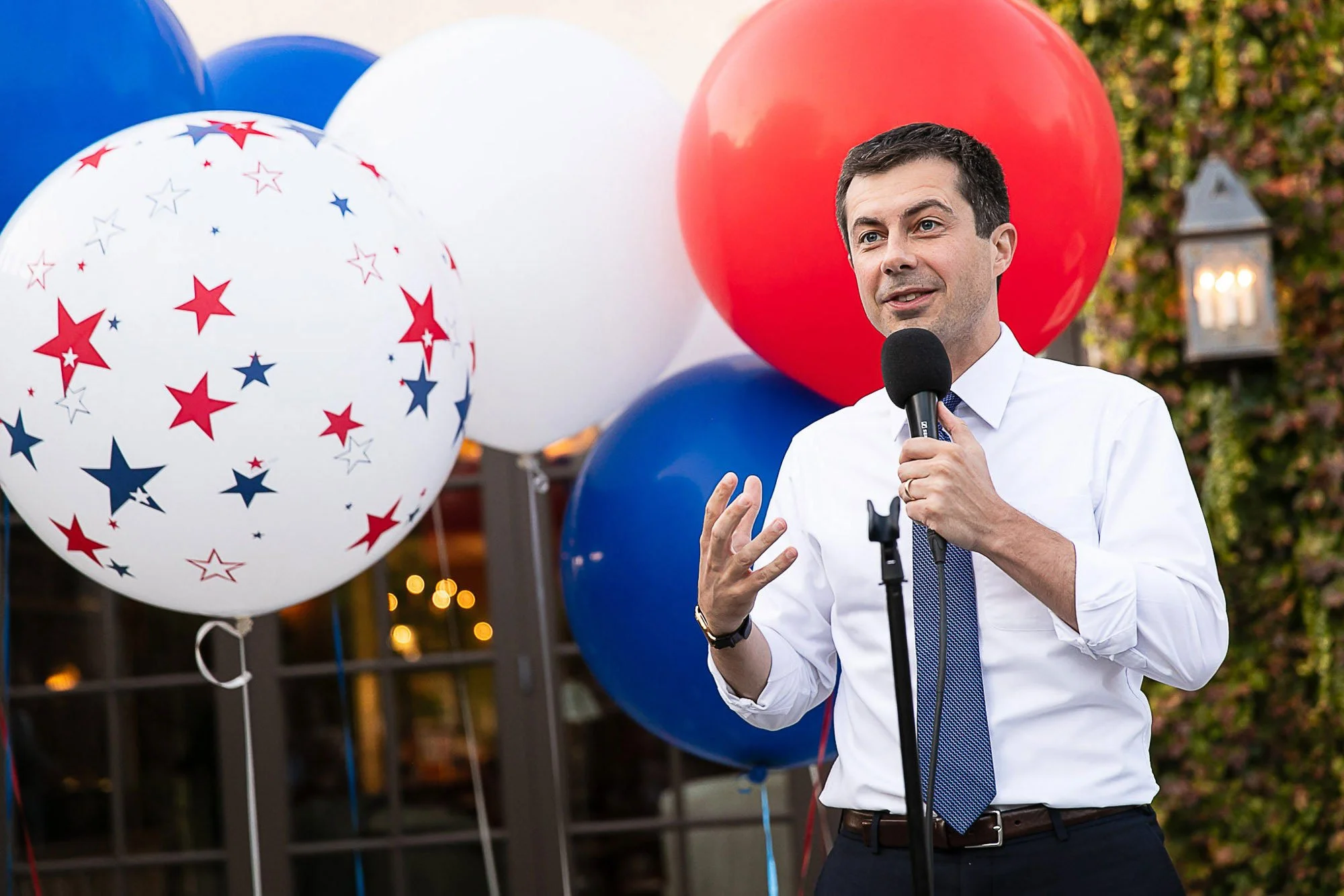 Pete Buttigieg speaks into a microphone at an outdoor event decorated with red, white, and blue balloons.