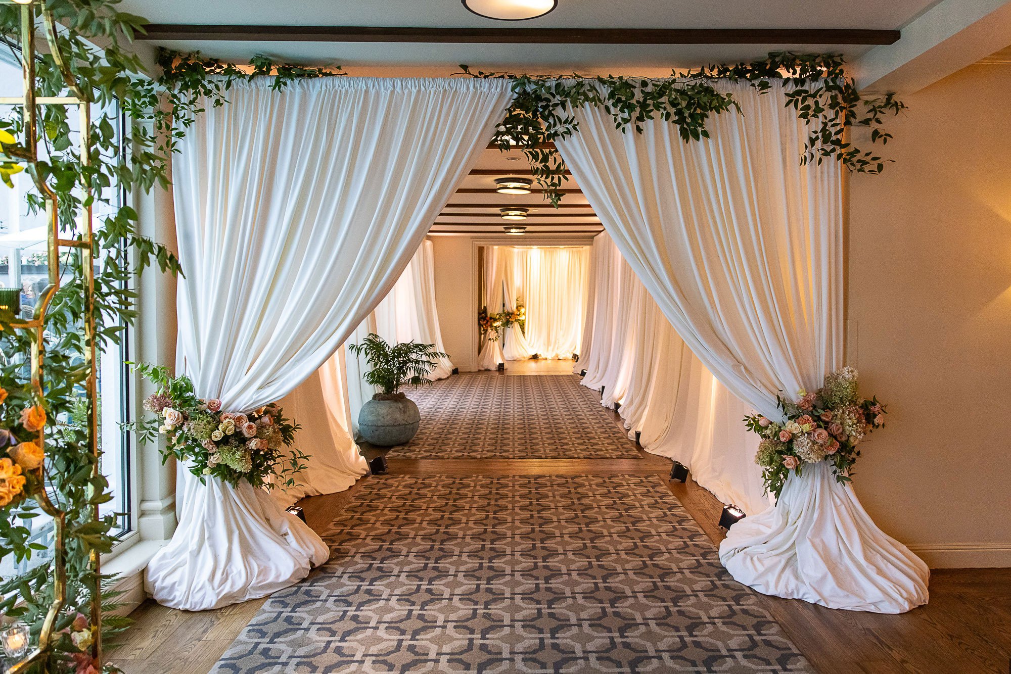 A hallway decorated with white curtains and floral arrangements, leading to an area with more decorations and flowers at the Beach and Tennis Club in Pebble Beach, CA.