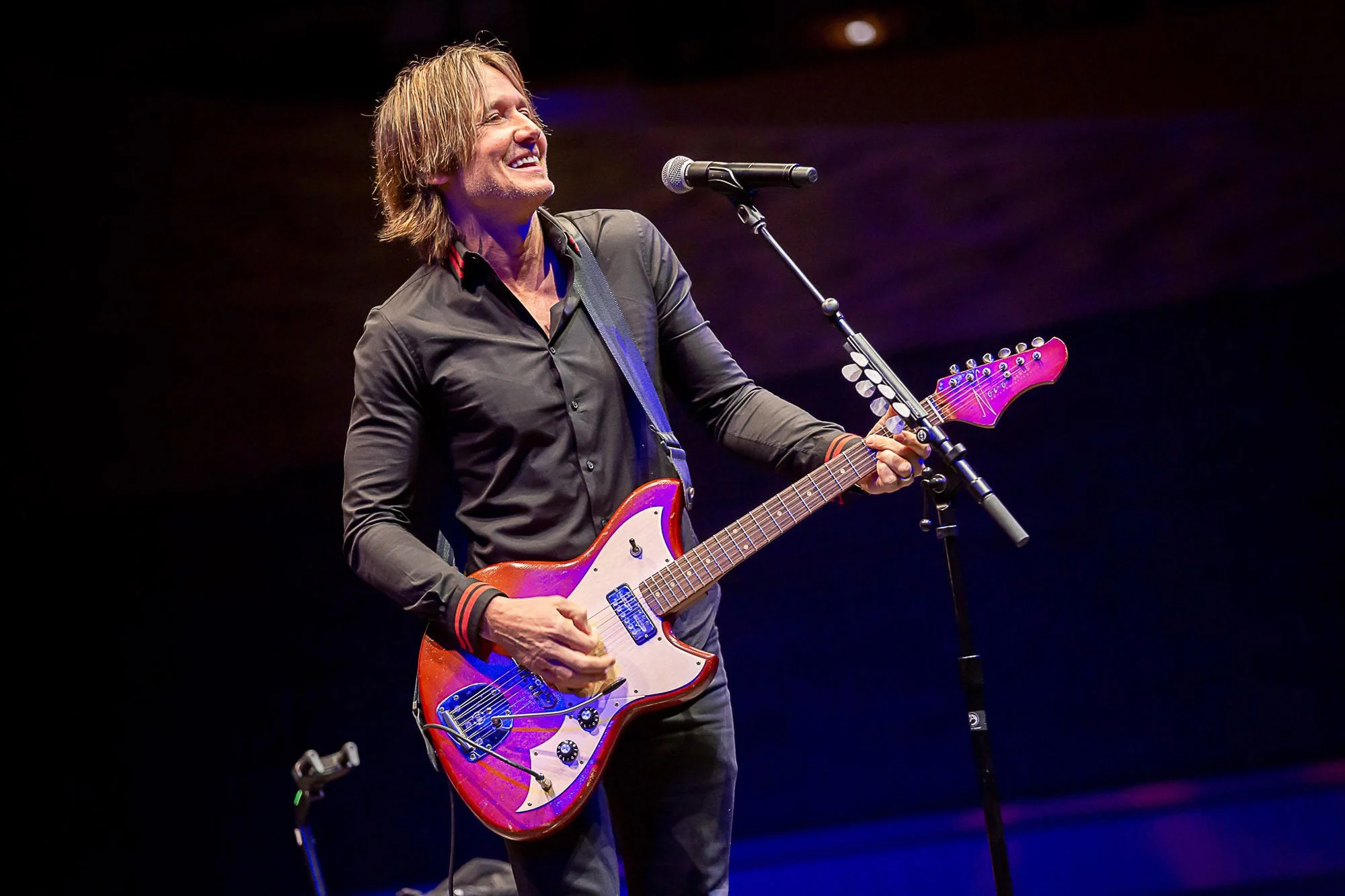 Keith Urban with shoulder-length blonde hair playing a pink electric guitar on stage with a dark background.