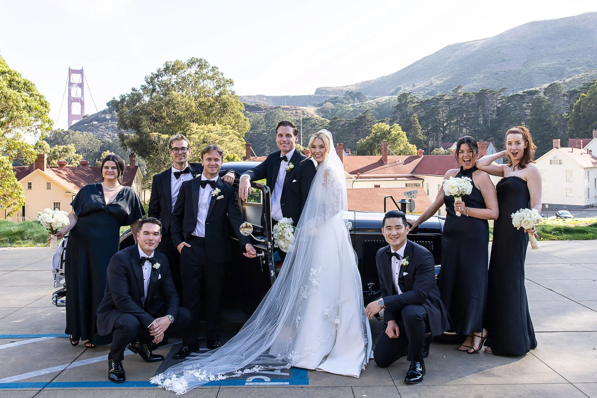 Group of wedding guests, including bride and groom, posing in front of a vintage car outdoors with trees and hills in the background on a sunny day at Cavallo Point in Sausalito, CA.