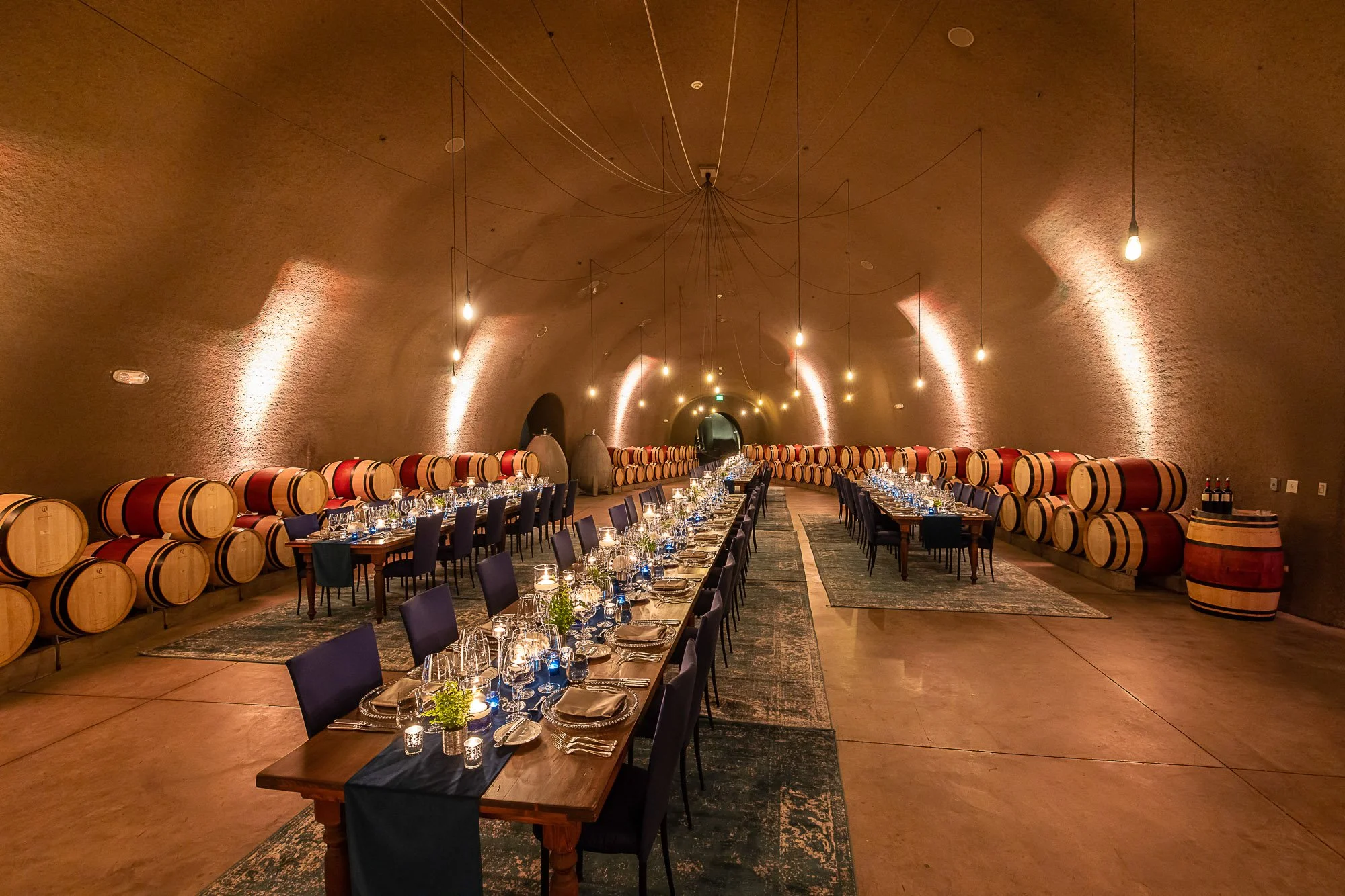Long dining table set for a formal event in a large wine cellar with wine barrels along the walls, warm lighting, and elegant table settings.