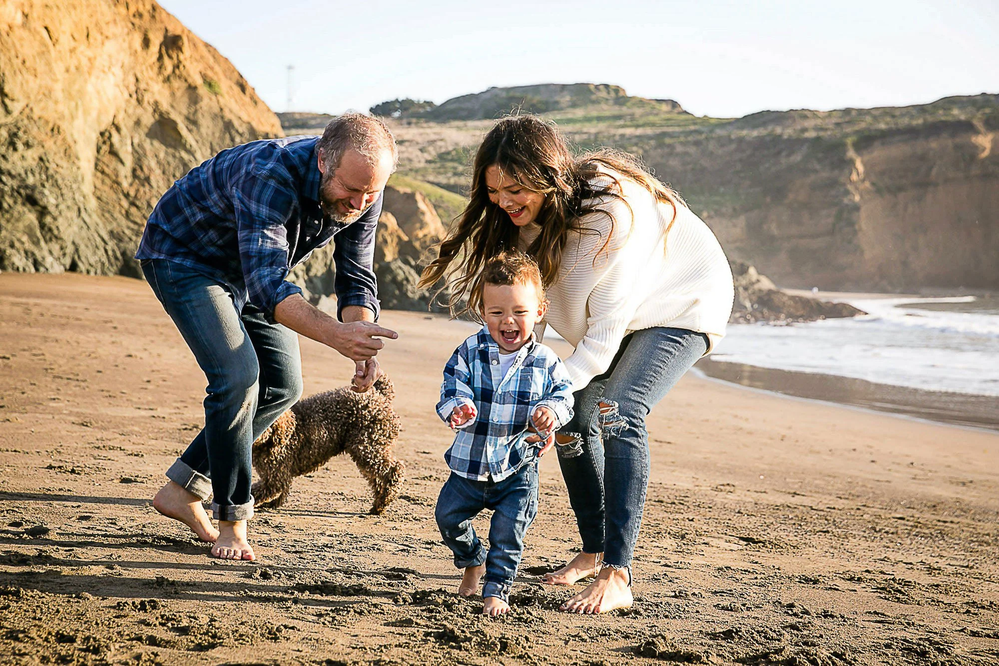 Family of three with a dog playing on the beach, with cliffs and the ocean in the background at Rodeo Beach in Marin California.
