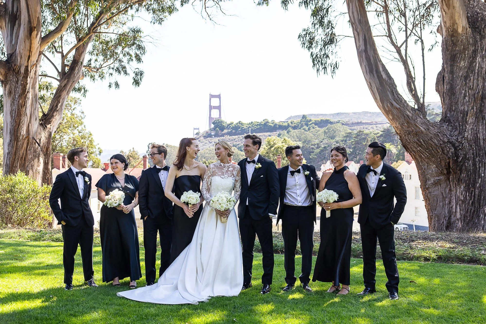 Group of wedding guests, including bride in white lace wedding gown and groom in black tuxedo, standing outdoors under trees with a view of the Golden Gate Bridge in the background at Cavallo Point in Sausalito, CA.