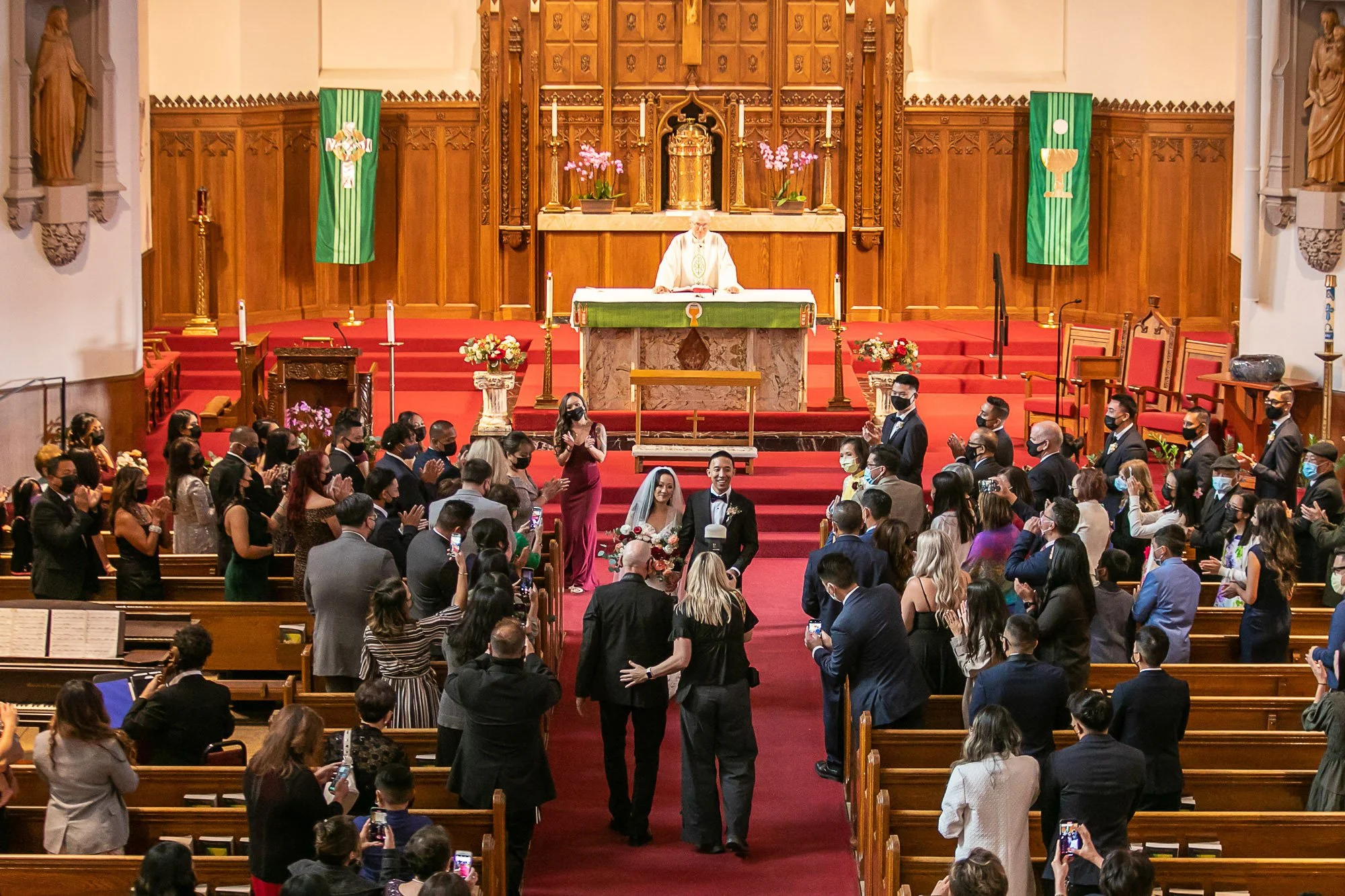 A church interior during a wedding ceremony, with a priest at the altar, the bride and groom walking down the aisle, and guests seated and standing. Tara Arrowood and Martin are shooting stills and video.