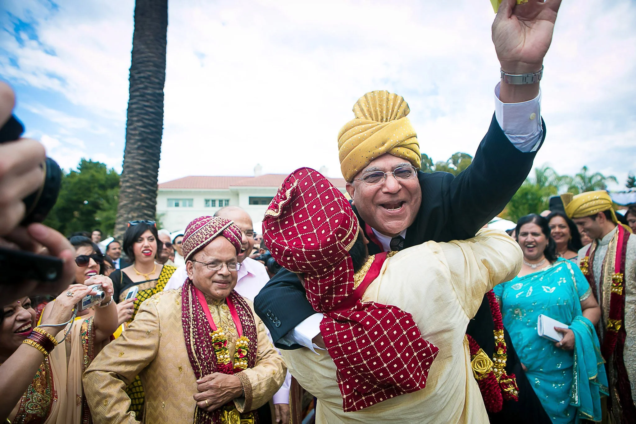 A group of people dressed in traditional Indian clothing, celebrating outdoors. A man in the center, wearing glasses and a yellow turban, is joyfully riding on the shoulders of a man in a patterned red and yellow turban. The crowd around them is smil