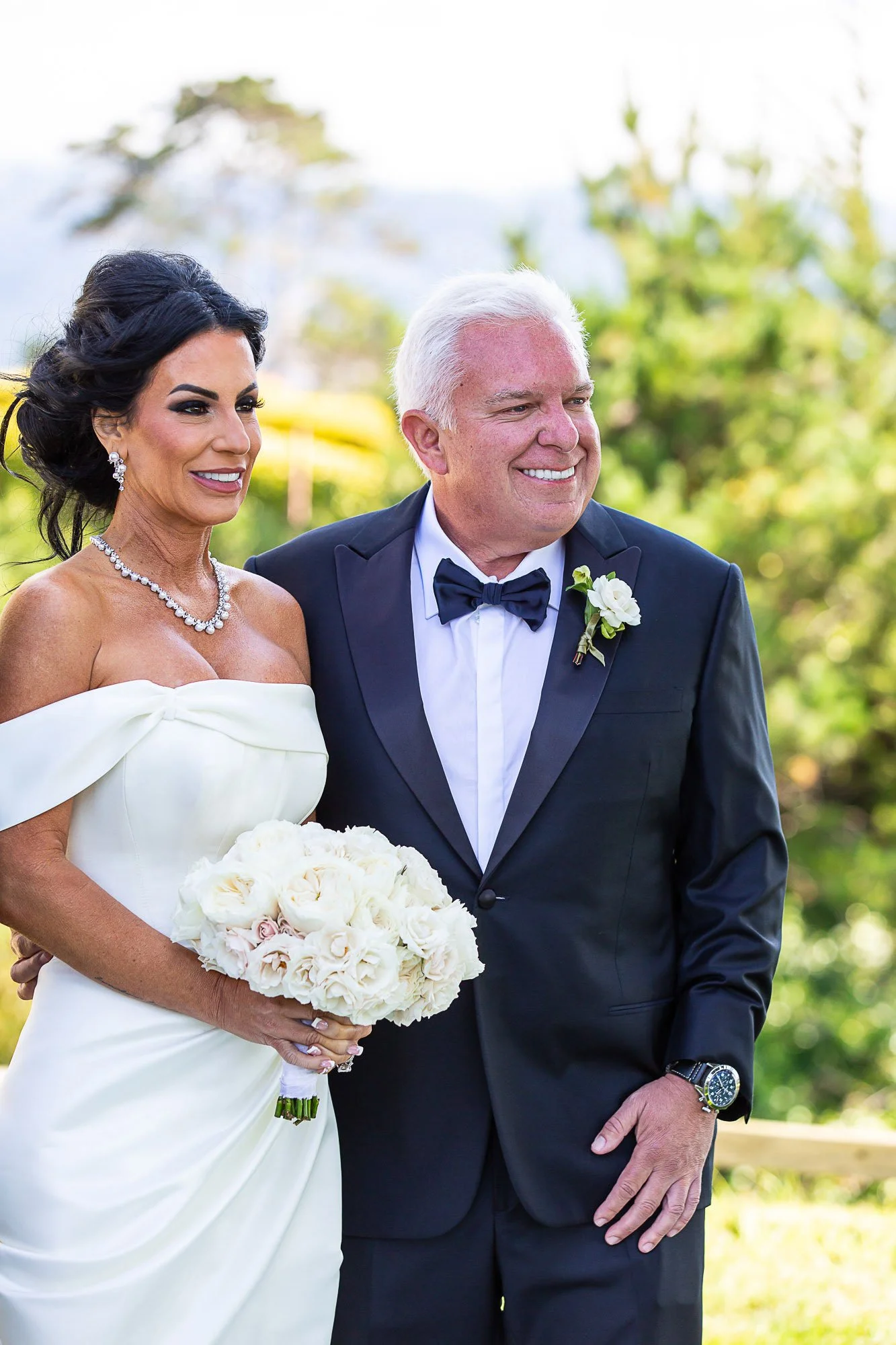 A bride and groom dressed in wedding attire standing outdoors on a sunny day, with the bride holding a bouquet of white roses and the groom wearing a tuxedo with a bow tie and floral boutonniere at the Beach and Tennis Club in Pebble Beach, CA.