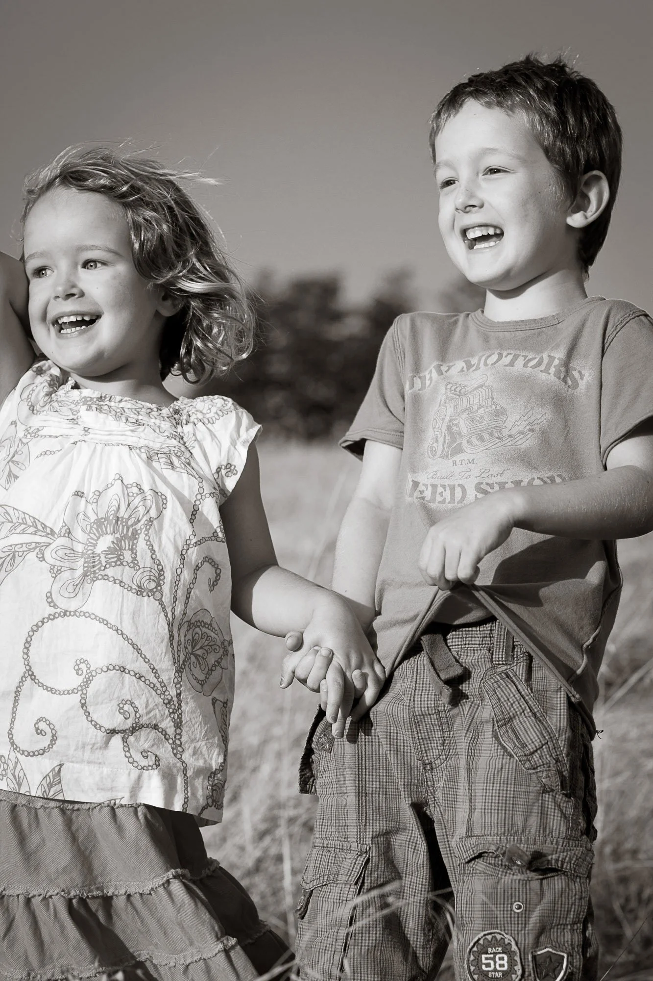 Two children, a girl with curly hair wearing a floral blouse and a boy with short hair wearing a graphic t-shirt, holding hands and smiling outdoors in a grassy area.