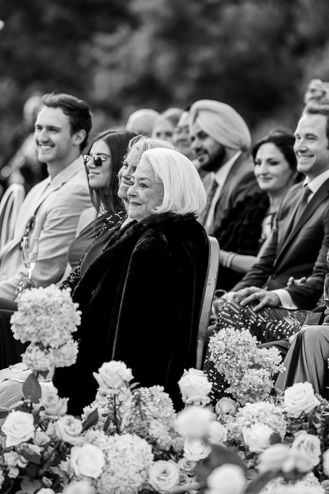 Black and white photograph of a group of people seated at the Beach and Tennis Club in Pebble Beach, smiling and dressed in formal attire, with floral arrangements in the foreground.