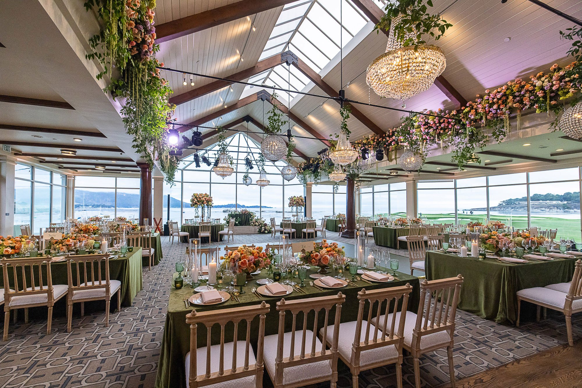 An elegant indoor reception area decorated with floral arrangements, hanging chandeliers, and scenic ocean views through large windows at the Beach and Tennis Club in Pebble Beach, CA.