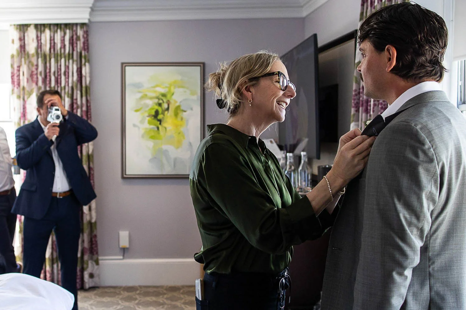 Tara Arrowood in glasses and a green blouse adjusting the tie of a man in a gray suit, smiling, in a well-decorated room with an abstract painting and floral curtains, with a man taking a photo in the background.