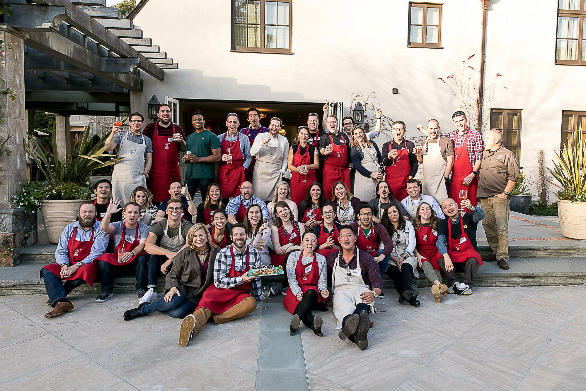 Group of diverse people gathered outside, smiling, and holding drinks, some wearing aprons and chef's hats, in front of a white building with large windows, plants, and outdoor furniture.