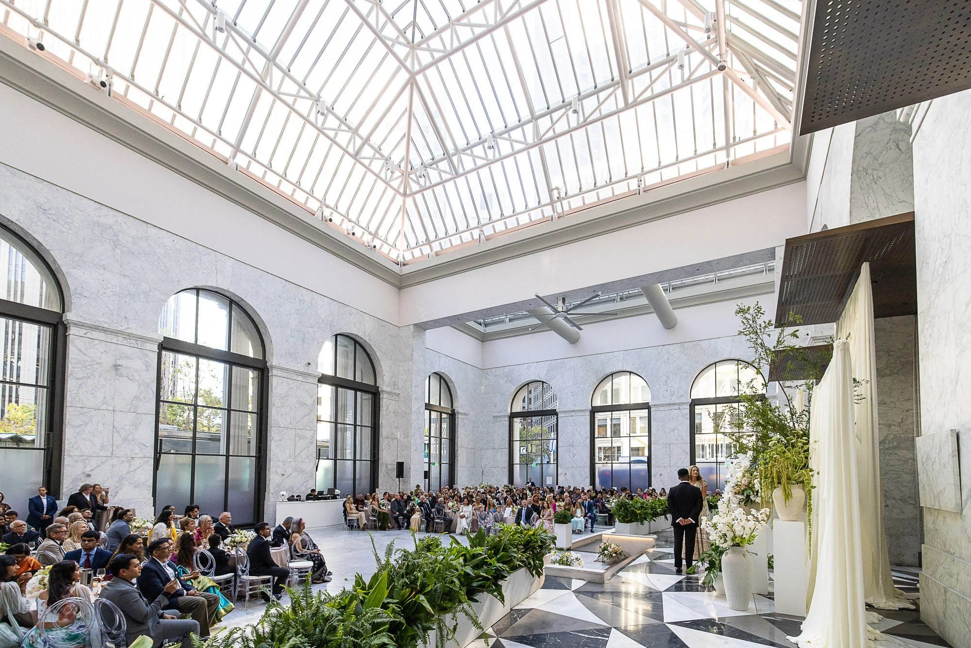 Multicultural Indian wedding ceremony beneath the glass atrium at the Conservatory at One Sansome in San Francisco, guests seated on the marble checkerboard floor