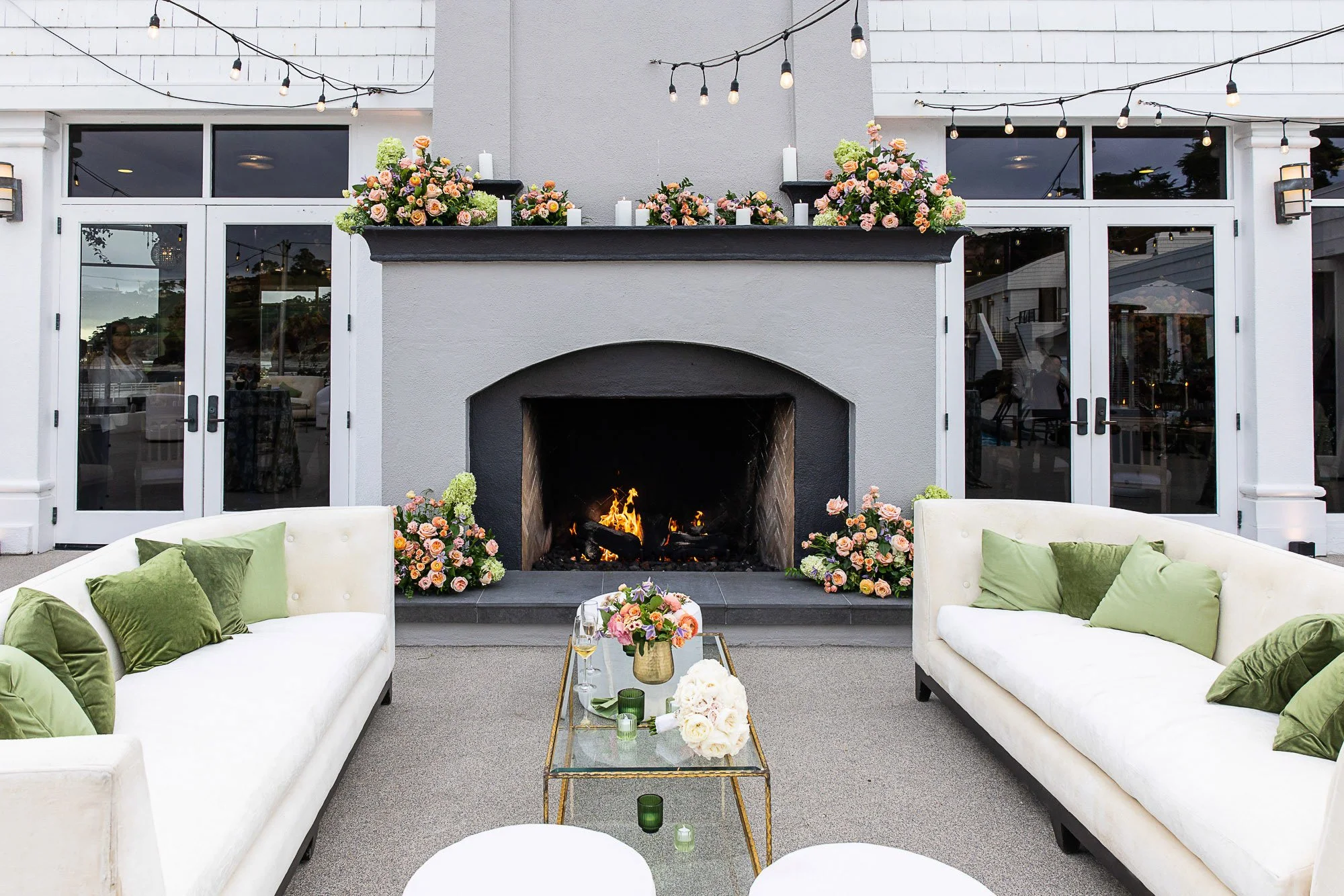 An outdoor patio with a fireplace, floral arrangements, string lights, and white sofas with green pillows at the Beach and Tennis Club in Pebble Beach, CA.