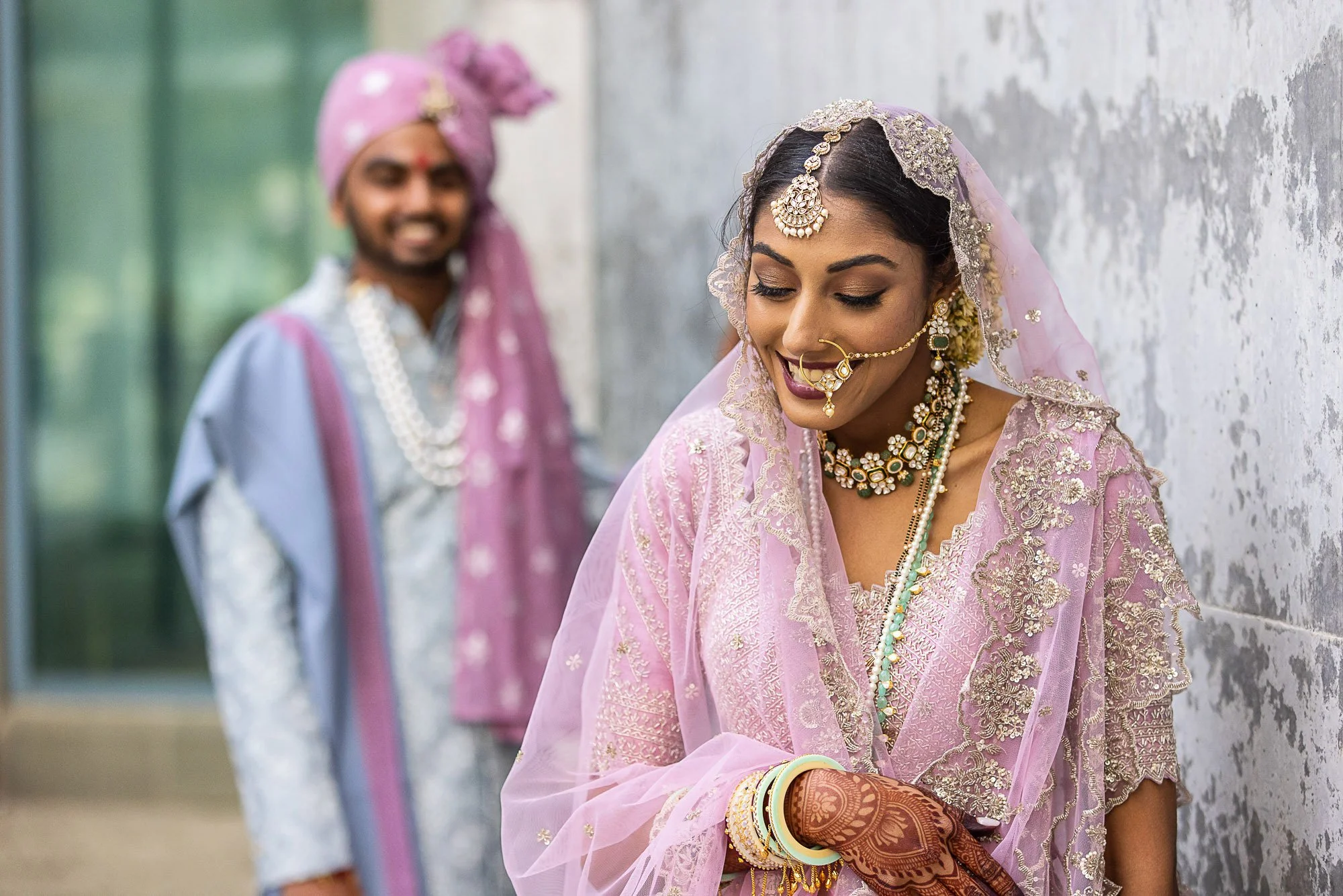 Indian bride wearing a pink and gold embroidered outfit with traditional jewelry, including a headpiece, nose ring, and bangles, smiling with her eyes closed, standing beside a groom dressed in traditional attire with a pink turban, against a texture