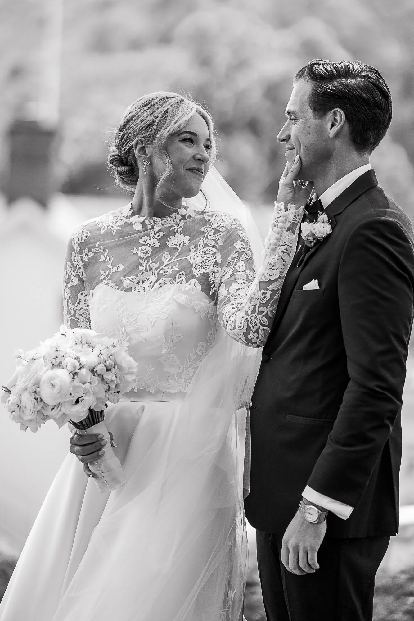 A black-and-white photograph of a bride and groom on their wedding day. The bride is holding a bouquet of flowers, wearing a lace wedding dress, and smiling at the groom. The groom is dressed in a dark suit with a boutonniere and looking at the bride