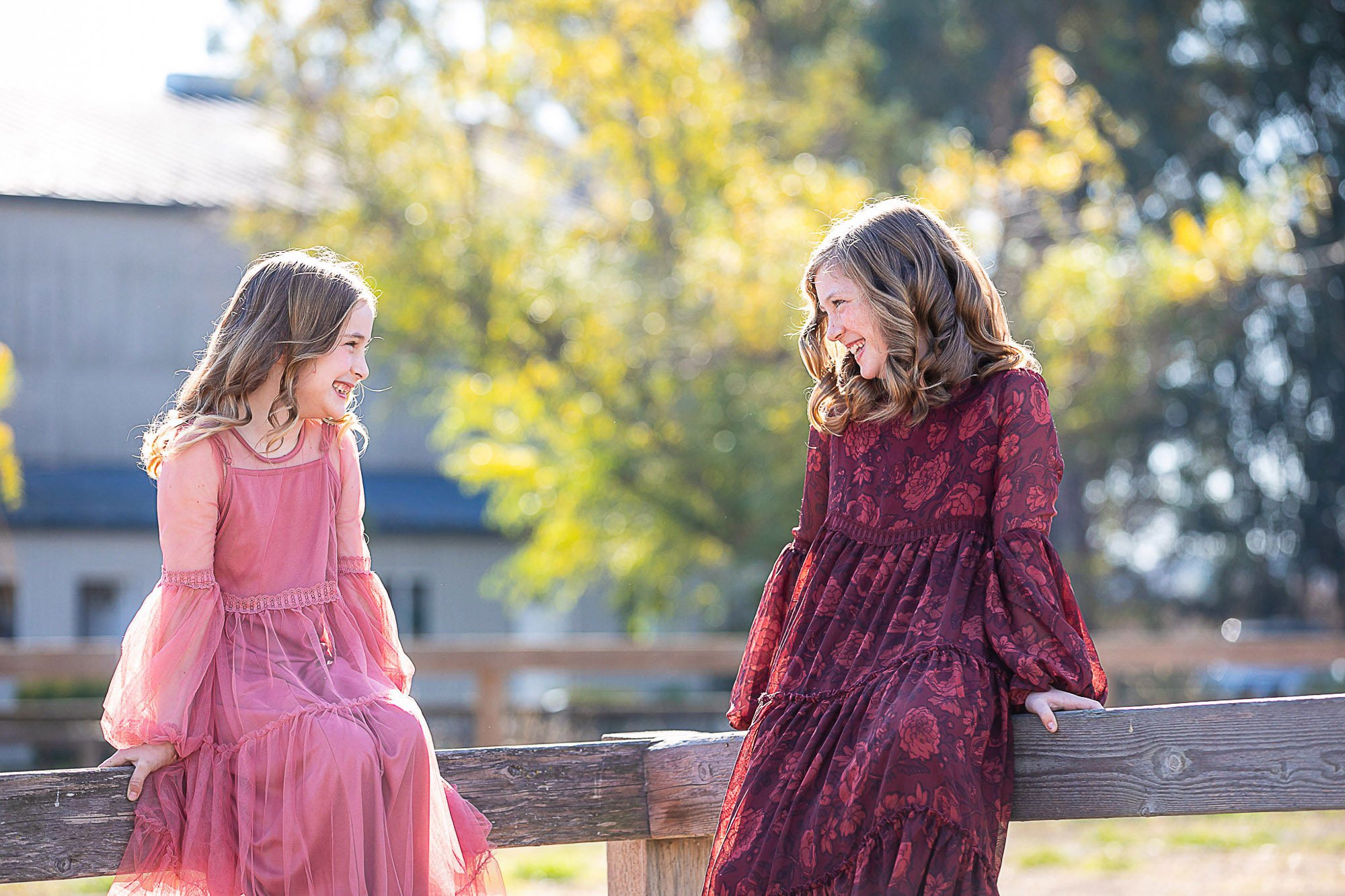 A woman and a girl sitting on a wooden fence outdoors, facing each other, smiling, and wearing long dresses, with trees and a house in the background.
