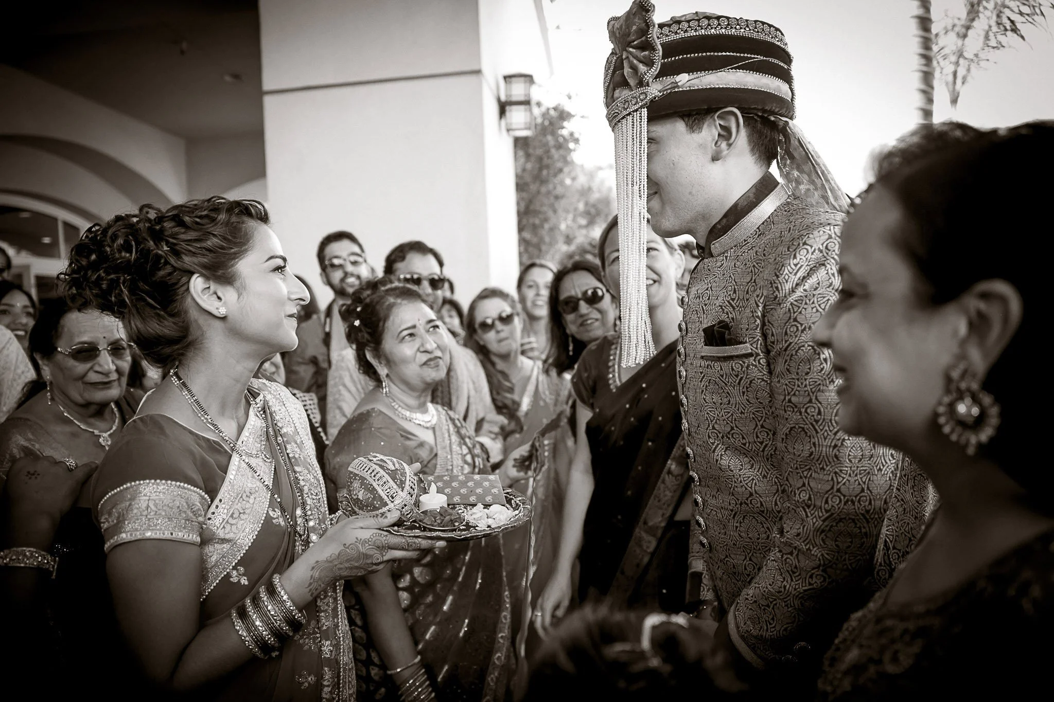 A traditional Indian wedding scene with women dressed in sarees, some wearing jewelry and bangles, gathered outdoors. The groom, wearing a richly embroidered sherwani and a turban with ornaments, is smiling and looking at a woman holding a tray with 