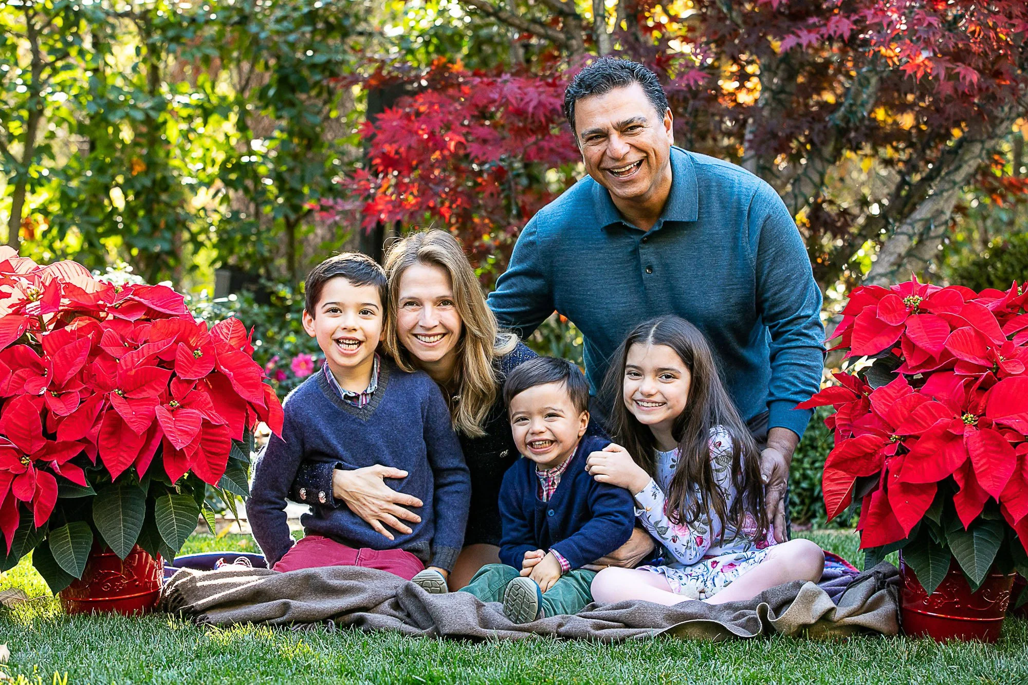 Family of six people, including three children and two adults, sitting on a blanket outdoors surrounded by red poinsettia plants and colorful trees, smiling for the camera.