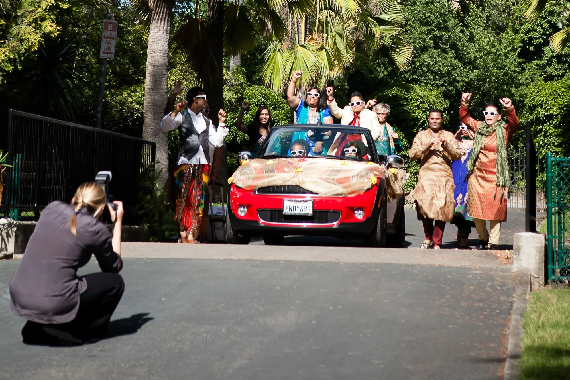 A group of people celebrating around a small red convertible car decorated with flowers and burlap, with some inside and some outside, holding their fists up, while a photographer kneels in front capturing the scene, set against a lush green backgrou