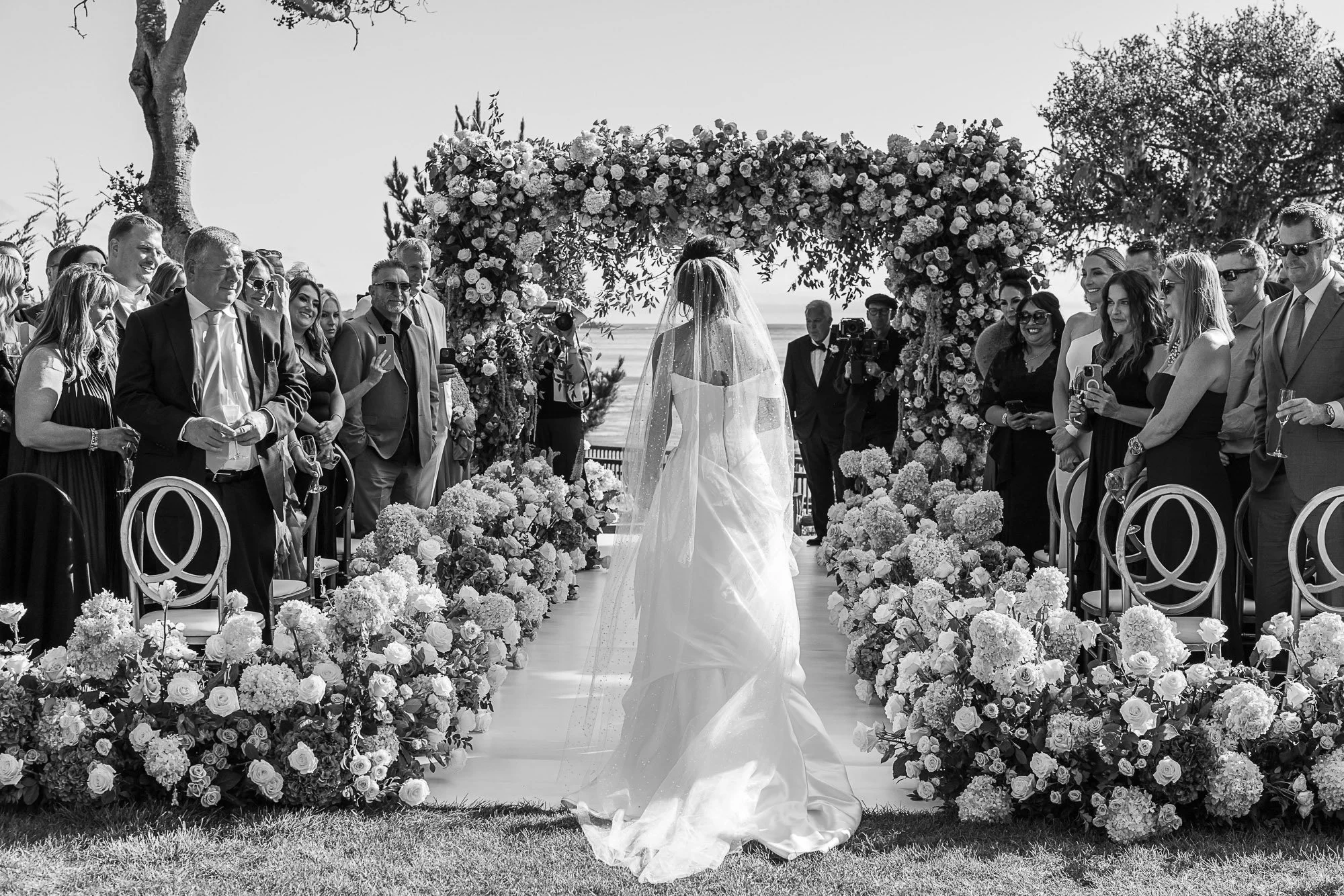 A black and white photo of a wedding ceremony outdoors with a bride standing under a decorated floral arch, surrounded by guests, with the ocean in the background at the Beach and Tennis Club in Pebble Beach, CA.