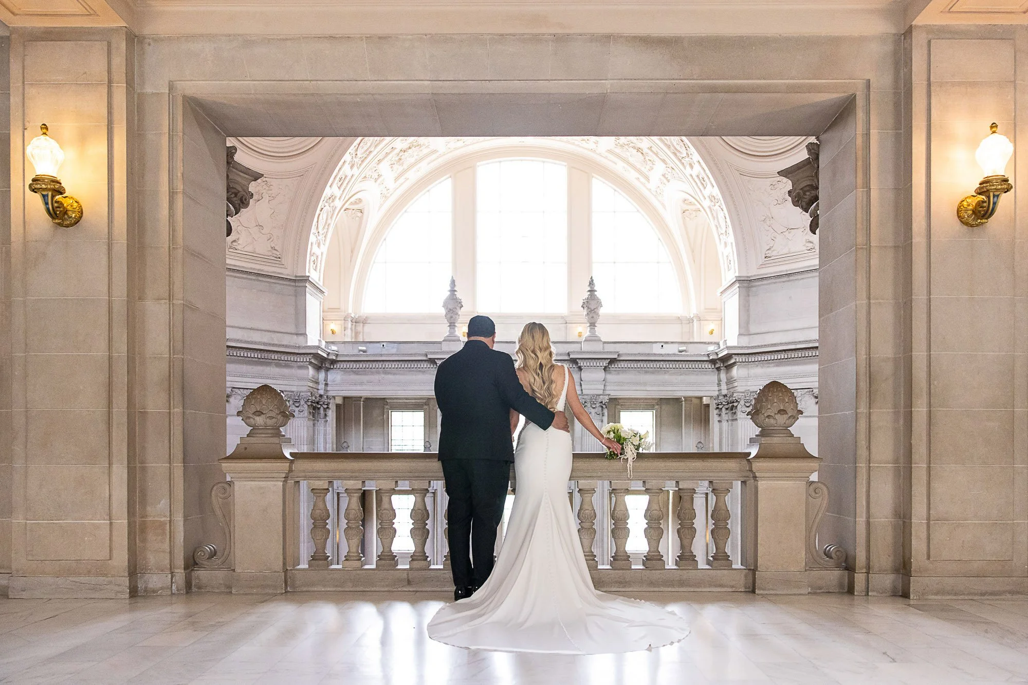 Bride and groom looking out over the marble balustrade beneath the arched window at San Francisco City Hall