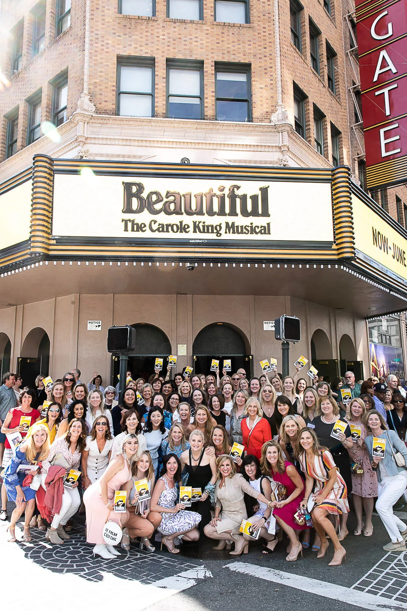 A large group of women gathered in front of a theater marquee for the musical 'Beautiful' - The Carole King Musical. They are smiling, holding up tickets or programs, and appear to be excited. The marquee indicates the show is now playing and runs th