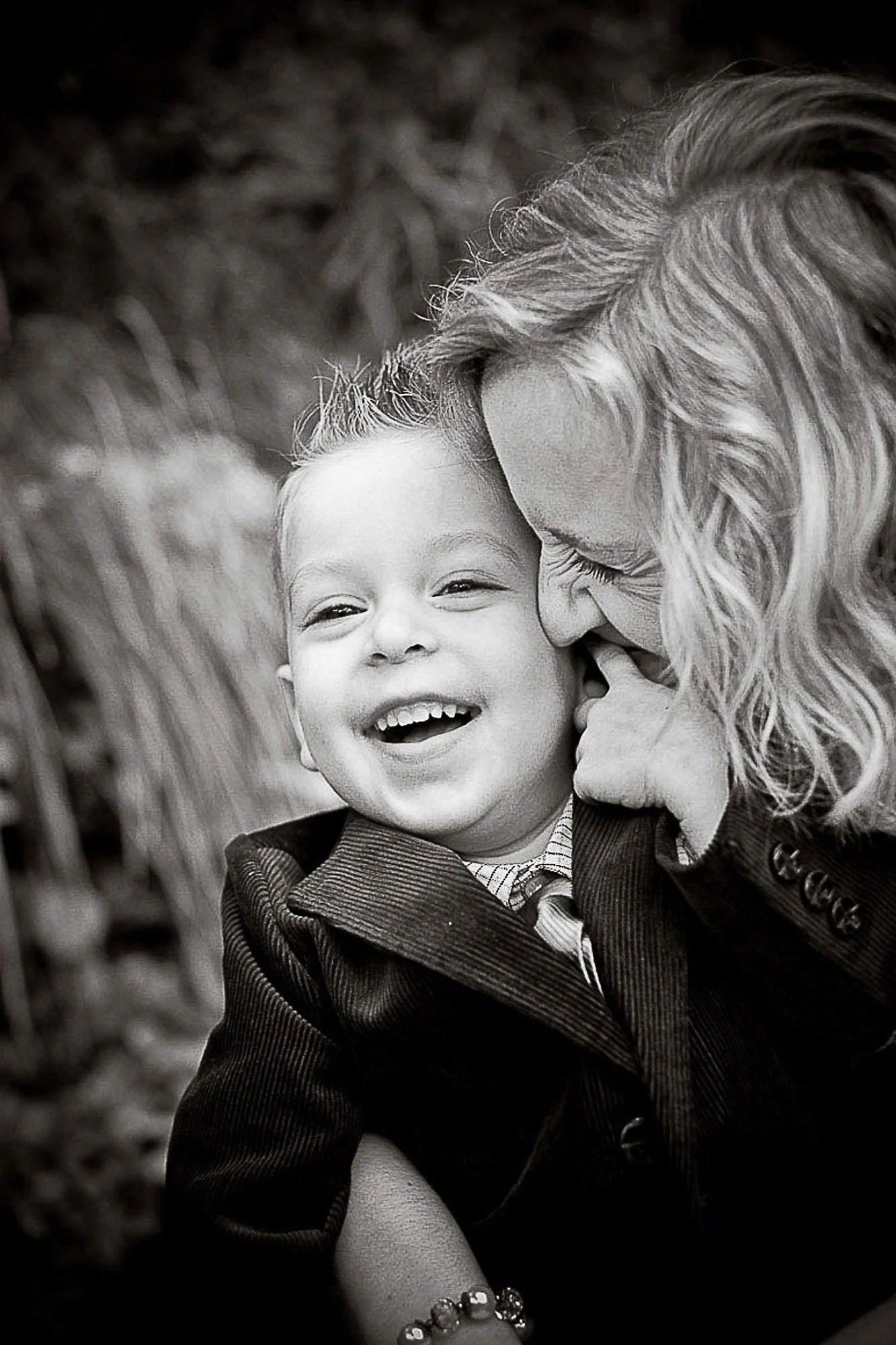 A woman with long, curly hair is cuddling a smiling young boy in a suit outdoors, both looking happy.