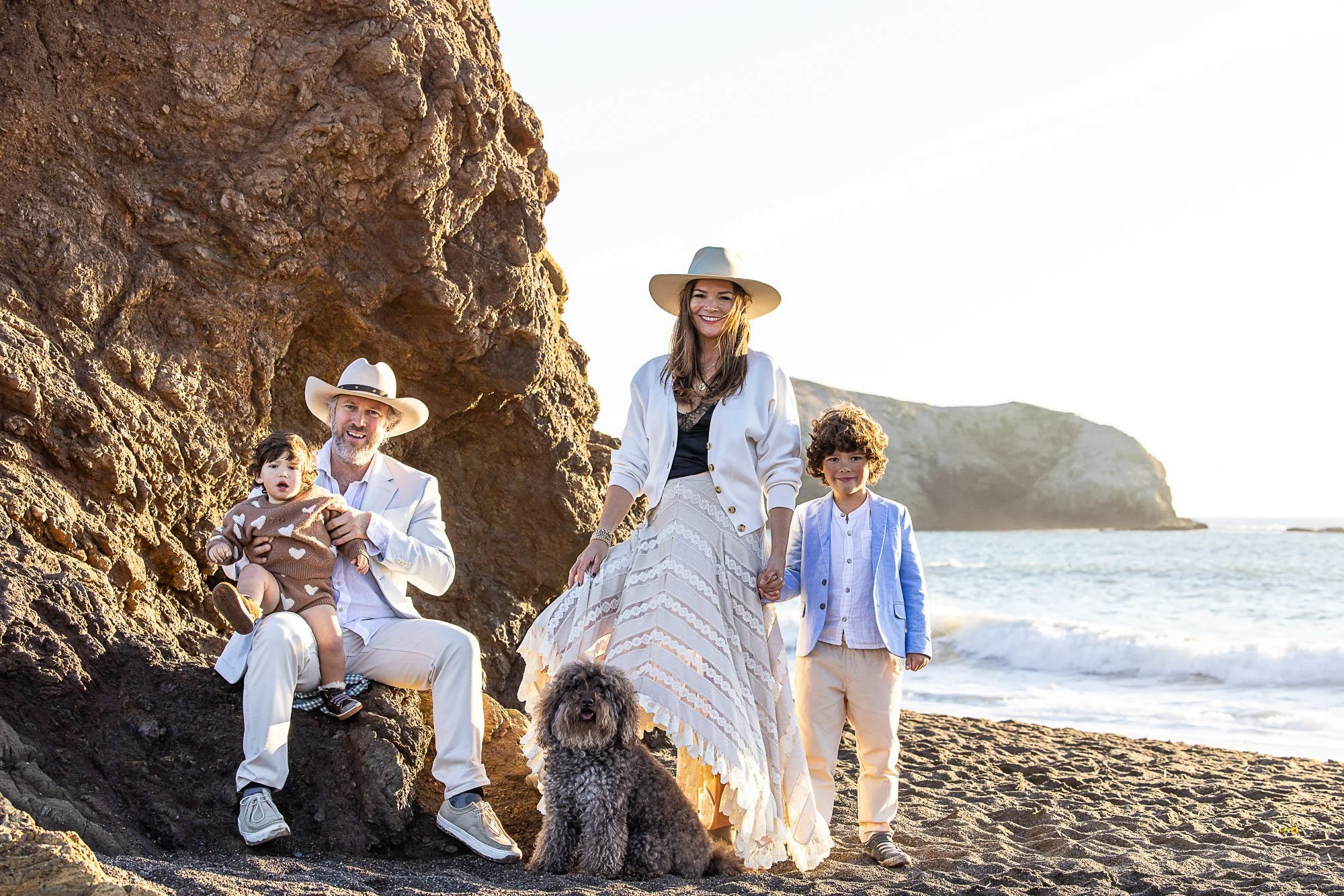 Family of five with a dog walking along a rocky beach during sunset. The family includes a woman, a man, two young boys, and a puppy, all dressed in light-colored clothing at Rodeo Beach in Marin California.