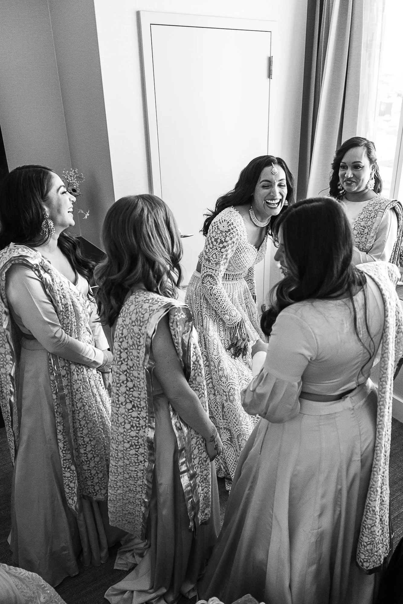 A group of women dressed in traditional Indian attire, chatting and laughing in a bright room with curtains and a window.