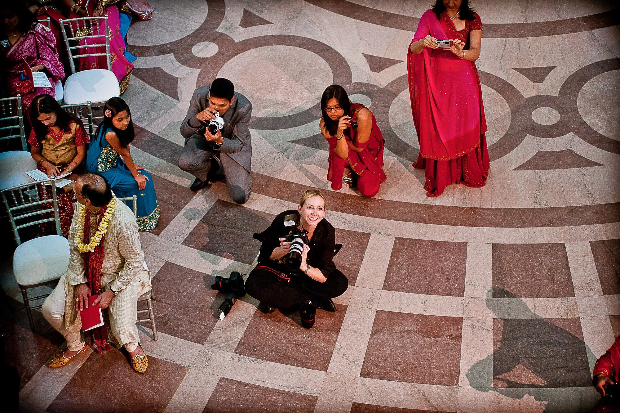 People dressed in colorful traditional Indian clothing, some seated and some standing, with Tara Arrowood taking photographs at City Hall with a decorative tiled floor.