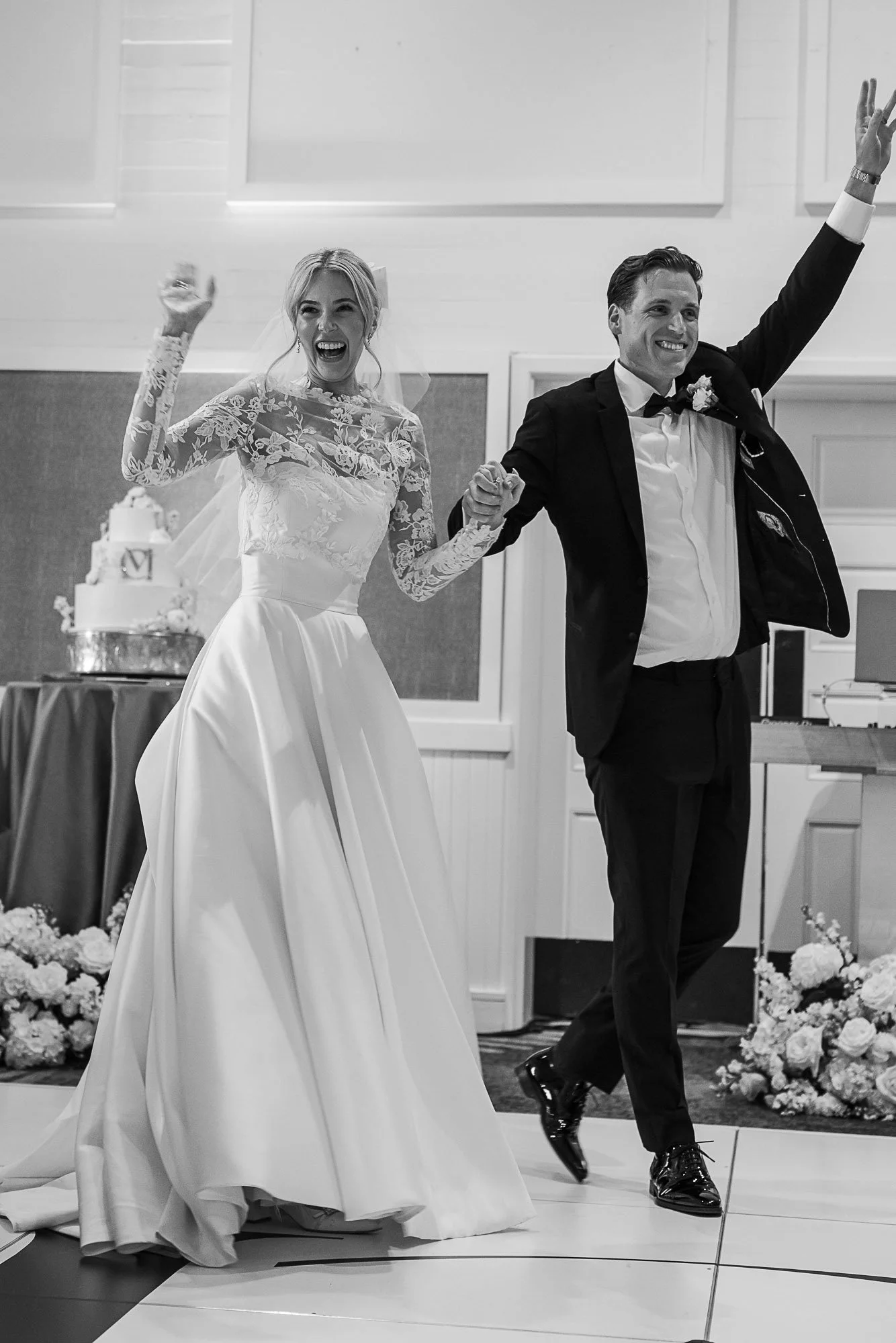 Black and white photo of a newlywed couple smiling and walking together at their wedding reception. The bride wears a lace wedding gown with long sleeves, and the groom is in a tuxedo with a boutonnière. They are holding hands and appear joyful, with