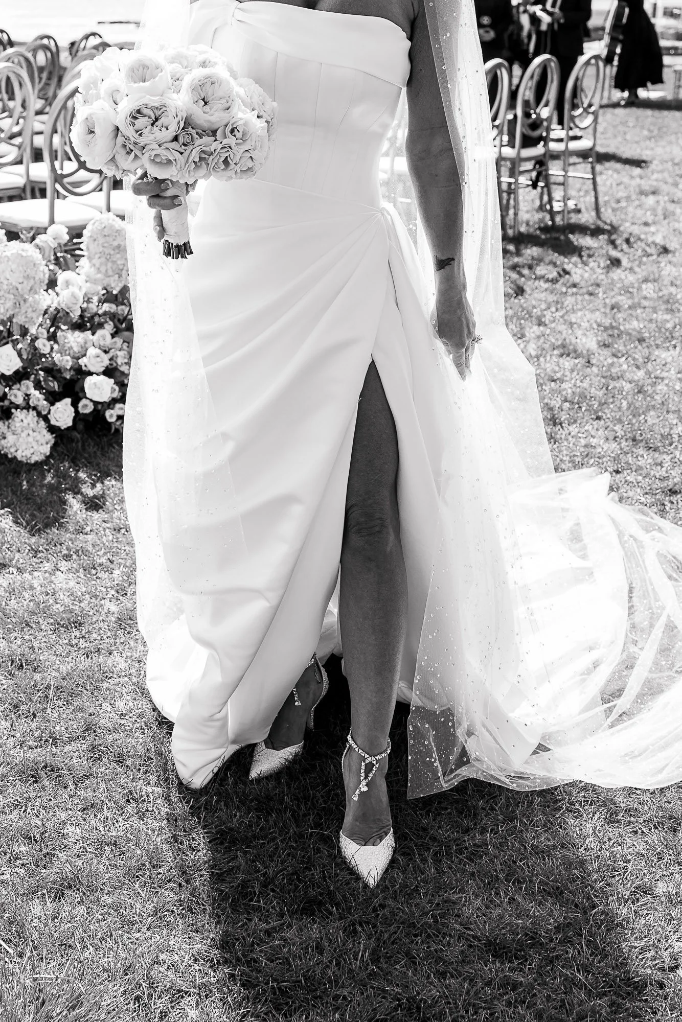 A bride in a wedding dress holding a bouquet, standing outdoors on grass, with chairs and flowers in the background at the Beach and Tennis Club in Pebble Beach, CA.