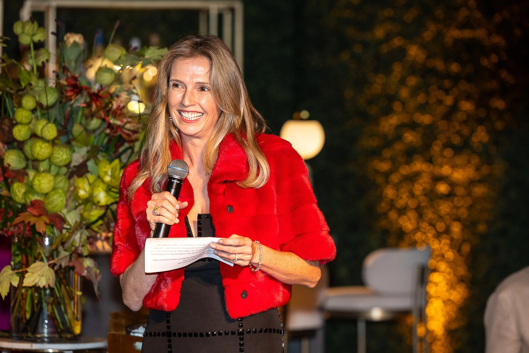 Woman with long blonde hair in a red jacket holding a microphone and reading from a paper at an outdoor event at night. There are yellow and orange lights in the background.