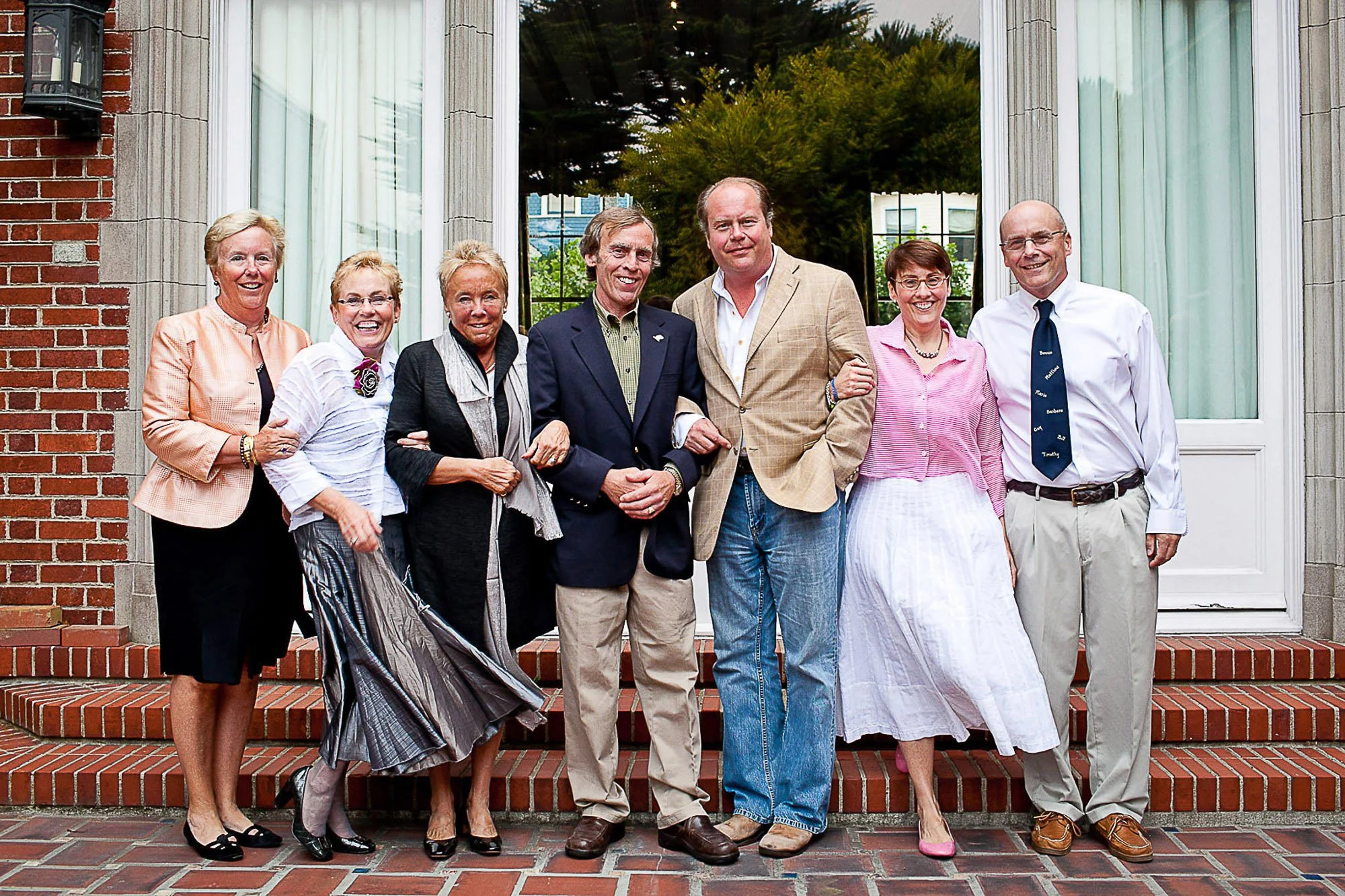 Group of seven adults, five women and two men, standing on brick steps outside a building with large windows and a door, smiling for the photo.