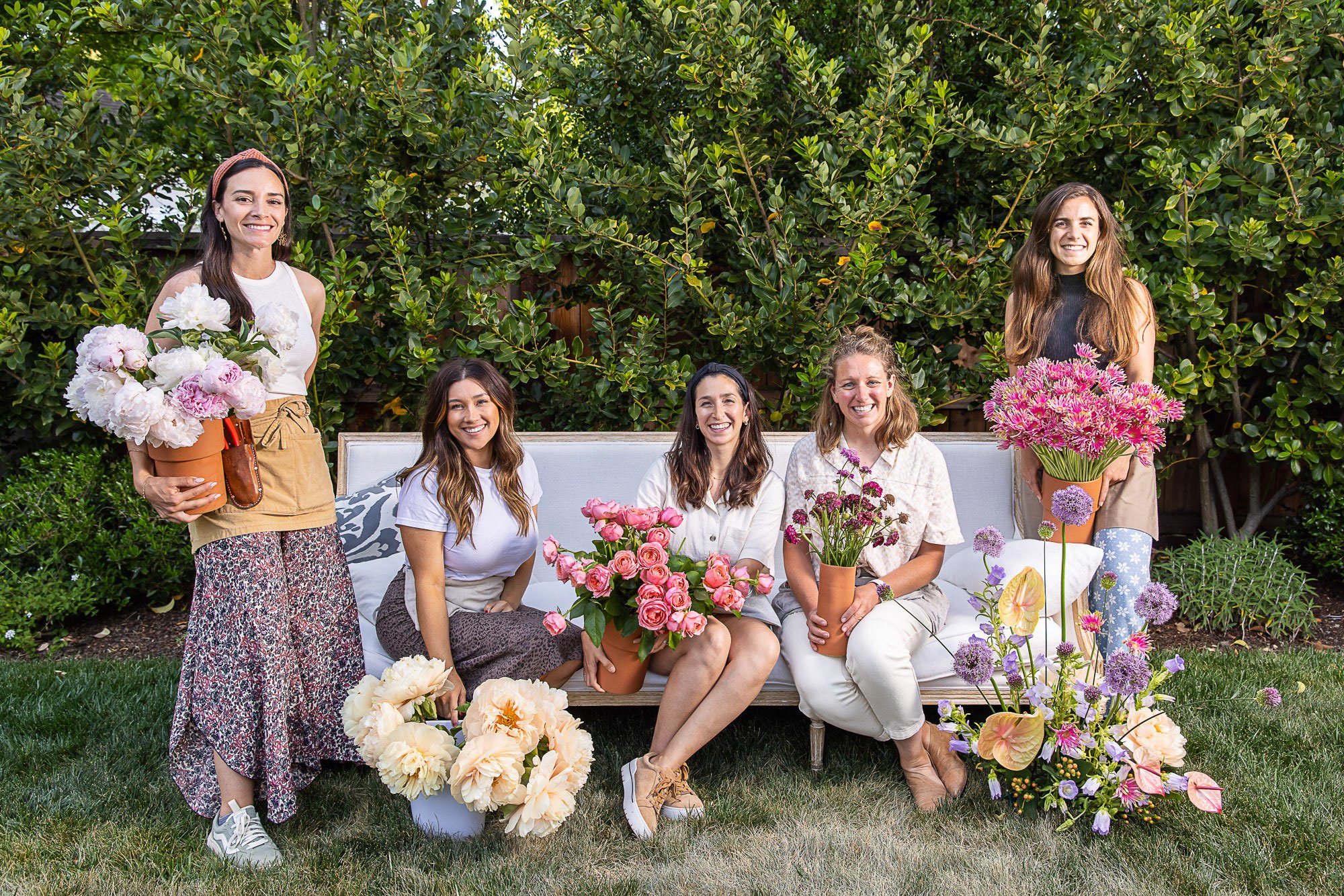 Six women are gathered outdoors in front of a large bush, sitting and standing on a grassy lawn, surrounded by numerous bouquets of colorful flowers. Some women are holding pots of flowers, and others have flowers on their laps or nearby, smiling at 