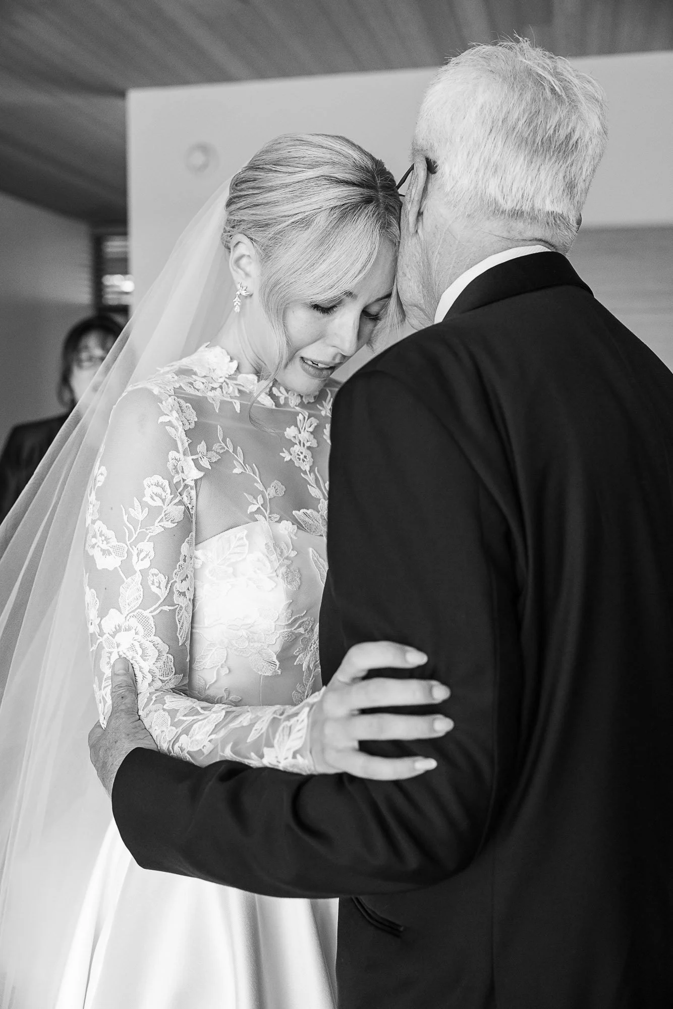 A black and white photo of a bride and an older man, possibly her father, embracing at her wedding. The woman, dressed in a lace wedding gown with a veil, has her eyes closed and a gentle expression. The older man, wearing a suit, holds her close wit