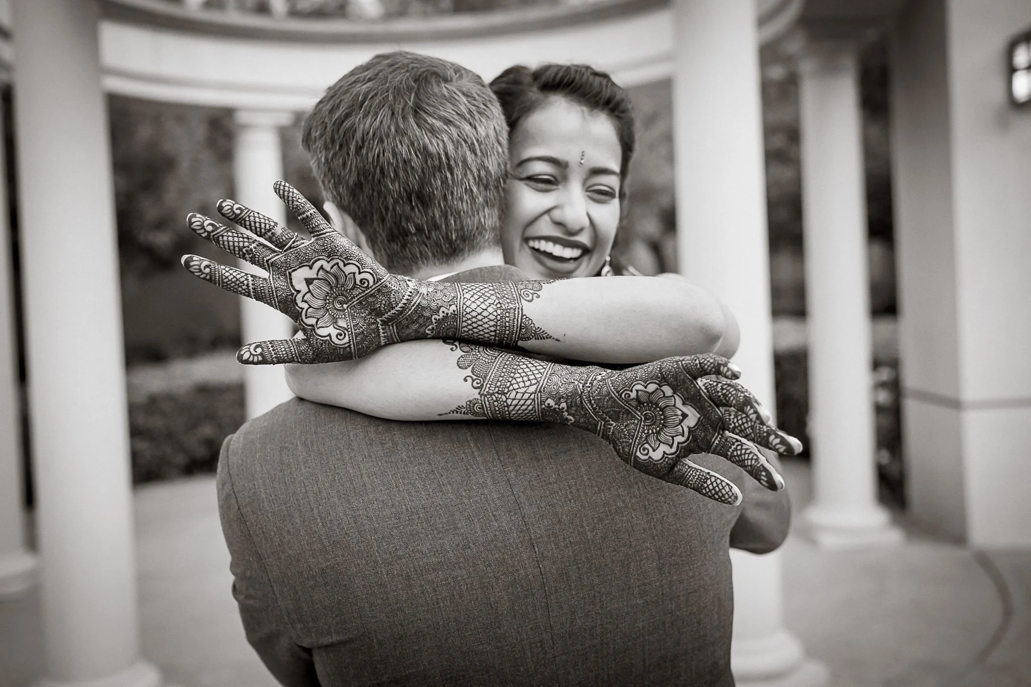 A couple hugs, with the woman smiling and showing henna on her hands, in an outdoor setting with columns in the background.