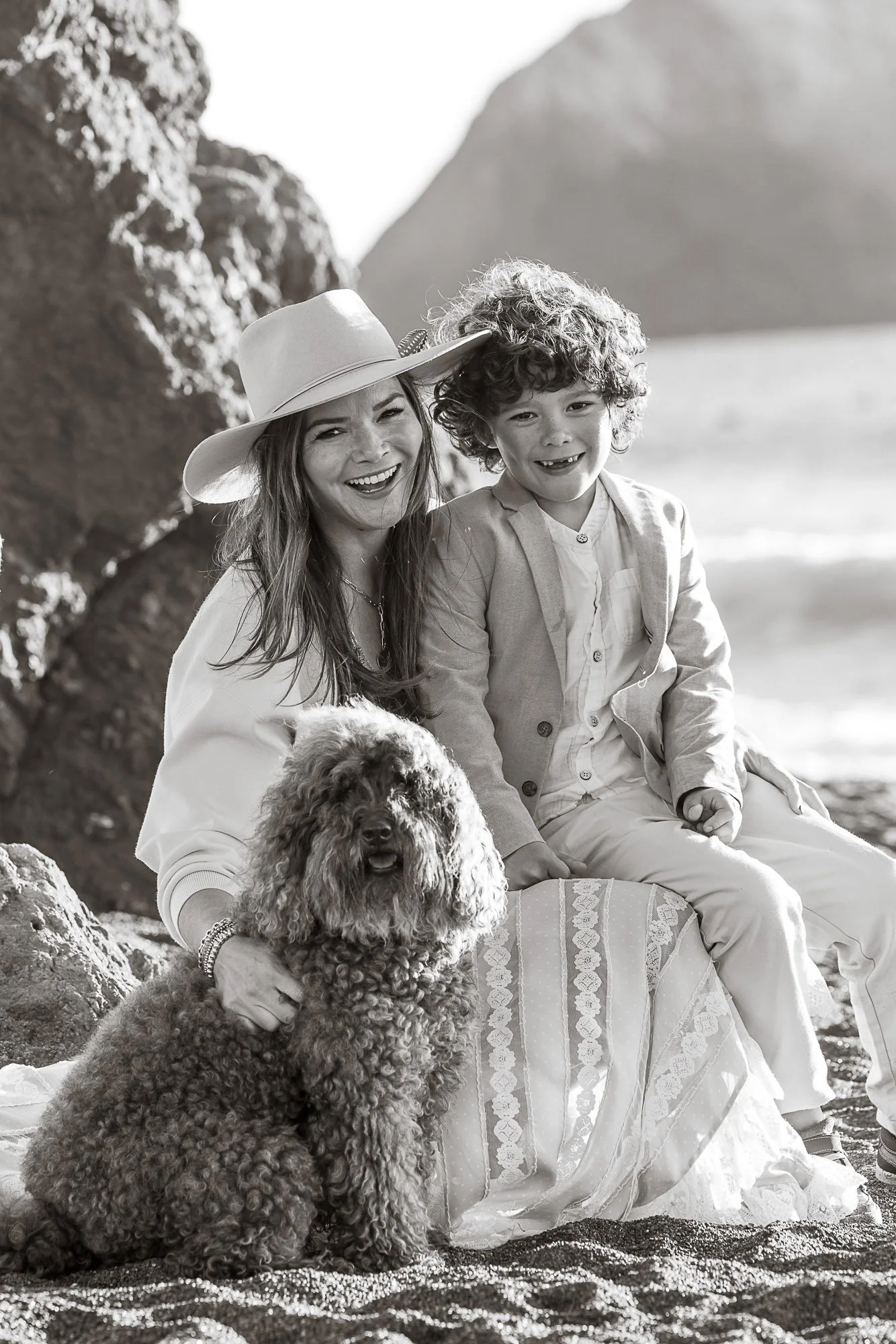 A woman and a young boy sitting on a blanket on a beach, with a large rock formation in the background. The woman is wearing a wide-brimmed hat and smiling, while the boy has curly hair and is also smiling. There are two dogs, one a large curly-haire