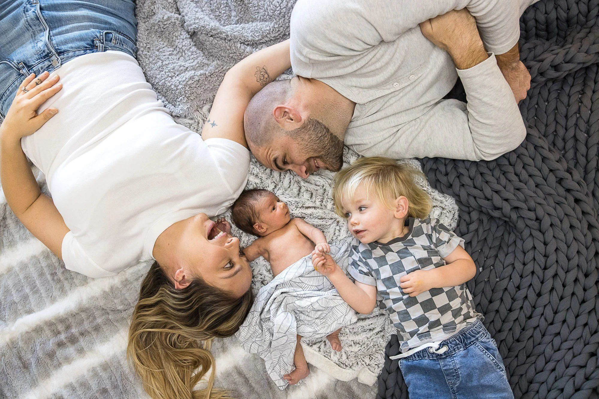 A woman, two young children, and two adult men lying on a plush rug, engaging in a playful and affectionate moment, with smiles and eye contact.