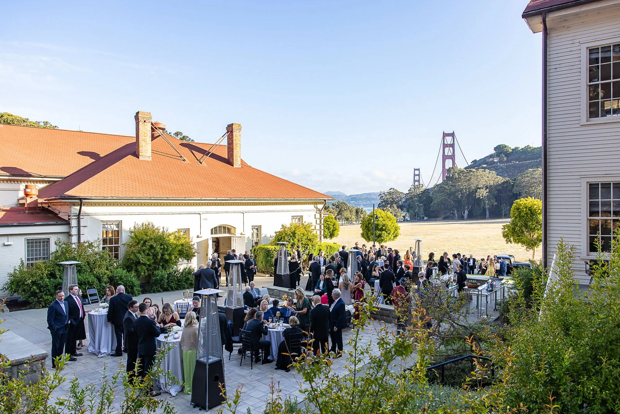 Outdoor wedding reception with many guests, tables, and heaters, set against a backdrop of the Golden Gate Bridge in San Francisco at Cavallo Point in Sausalito, CA.