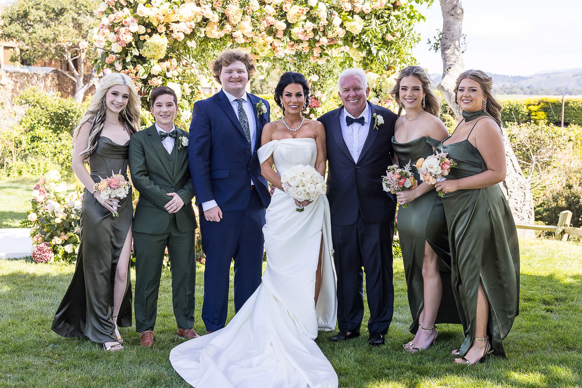 Group photo of six people, including a bride in a white gown and five others in formal attire, standing outdoors in front of a floral archway, with trees and a scenic background at the Beach and Tennis Club in Pebble Beach, CA.