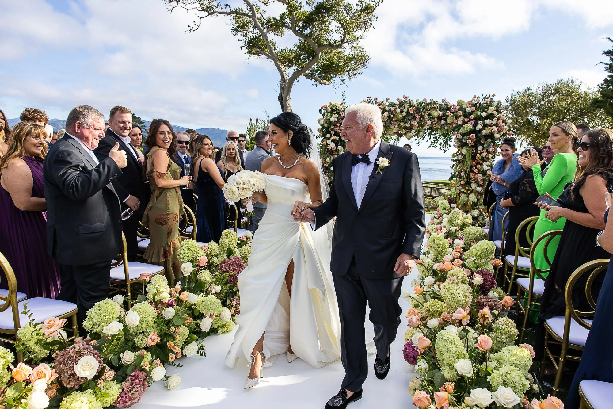 A bride and groom walking down the aisle at an outdoor wedding ceremony at the Beach and Tennis Club in Pebble Beach, surrounded by guests, with a floral arch and scenic seaside background.