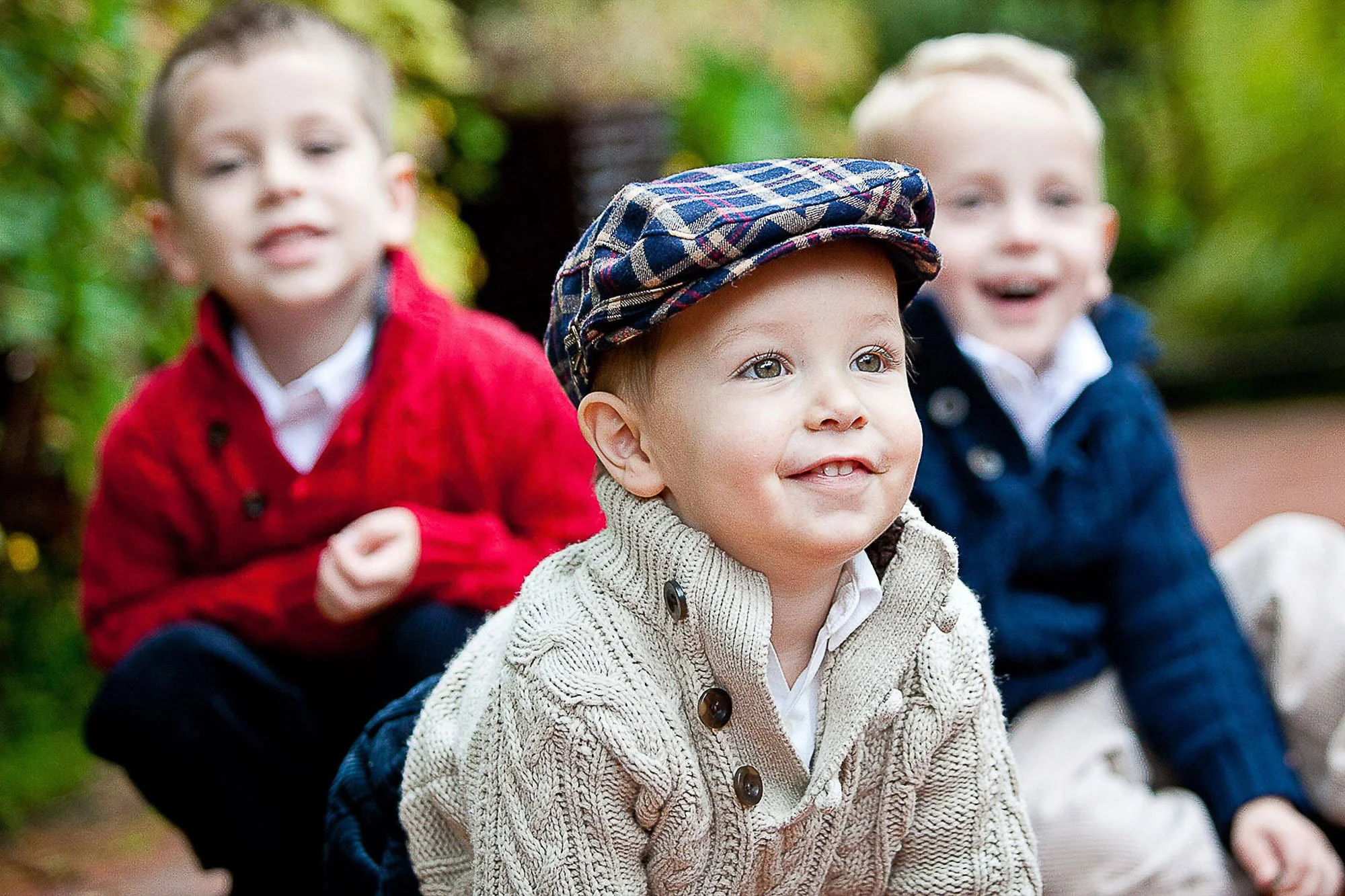 Three young children sitting outdoors on a park bench, smiling and enjoying the day. Two boys are in the background with blurred faces, and a young girl in the foreground wearing a beige knitted sweater and a checkered flat cap.