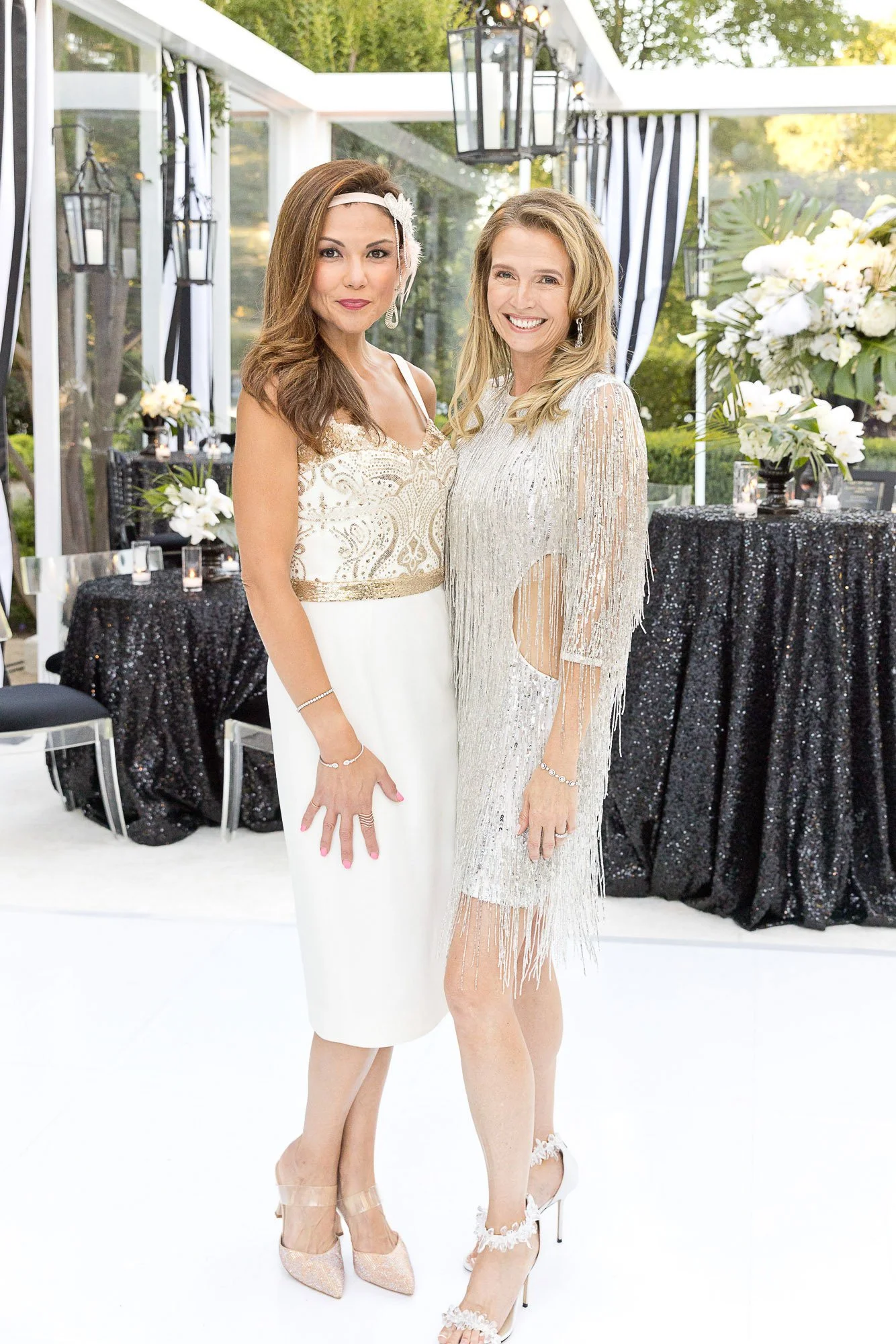 Two women dressed in elegant evening gowns smiling and posing together at an outdoor event, with decorated tables and greenery in the background.