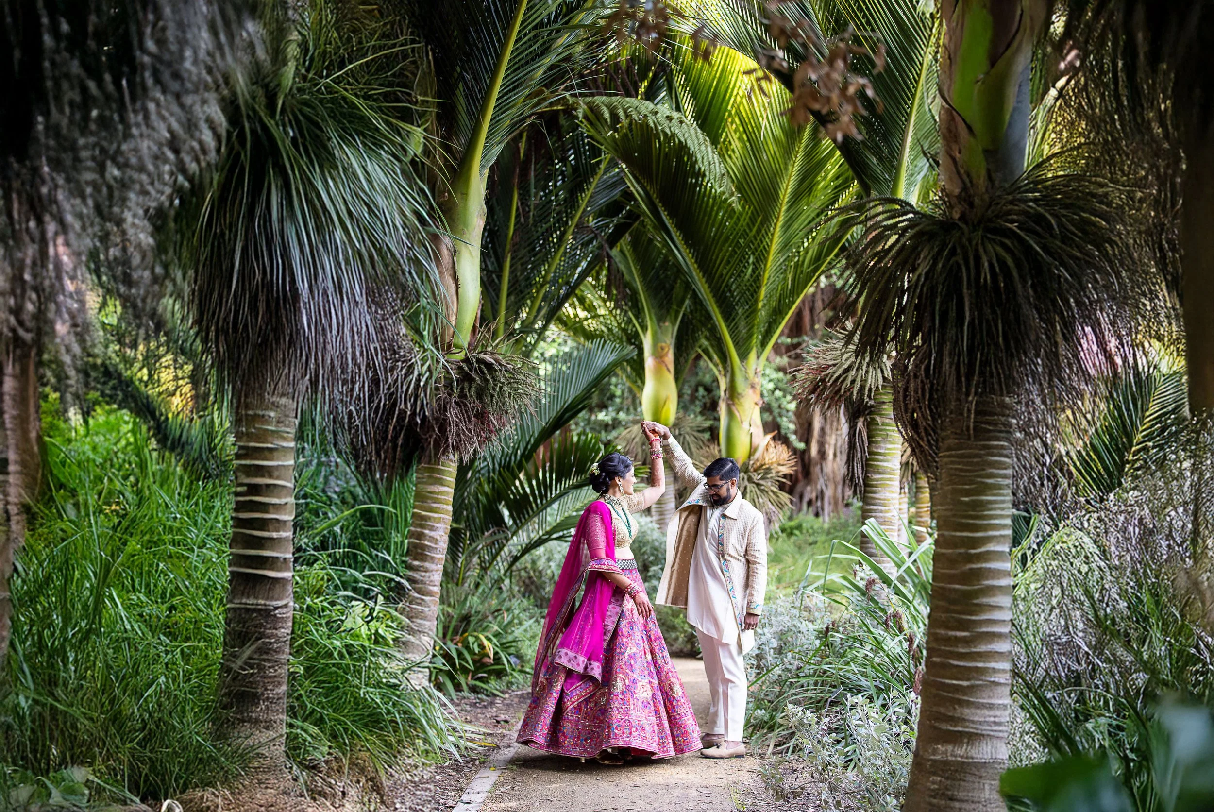 An Indian couple dressed in traditional Indian attire walking through a lush, green garden filled with tall palm trees at Botanical Gardens.