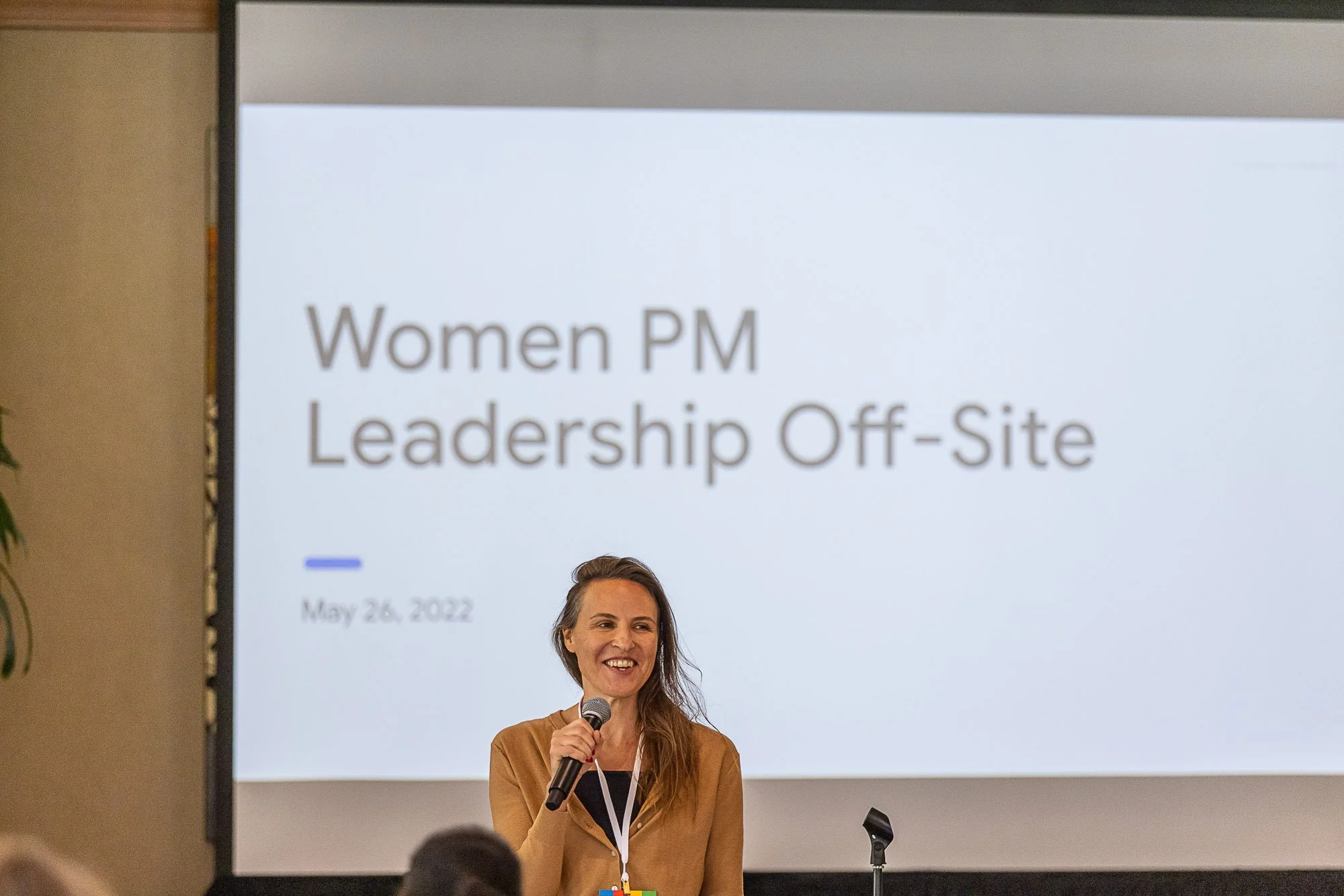A woman holding a microphone and smiling during a presentation at an event labeled 'Women PM Leadership Off-Site' on May 26, 2022, with a large screen behind her showing the event details.