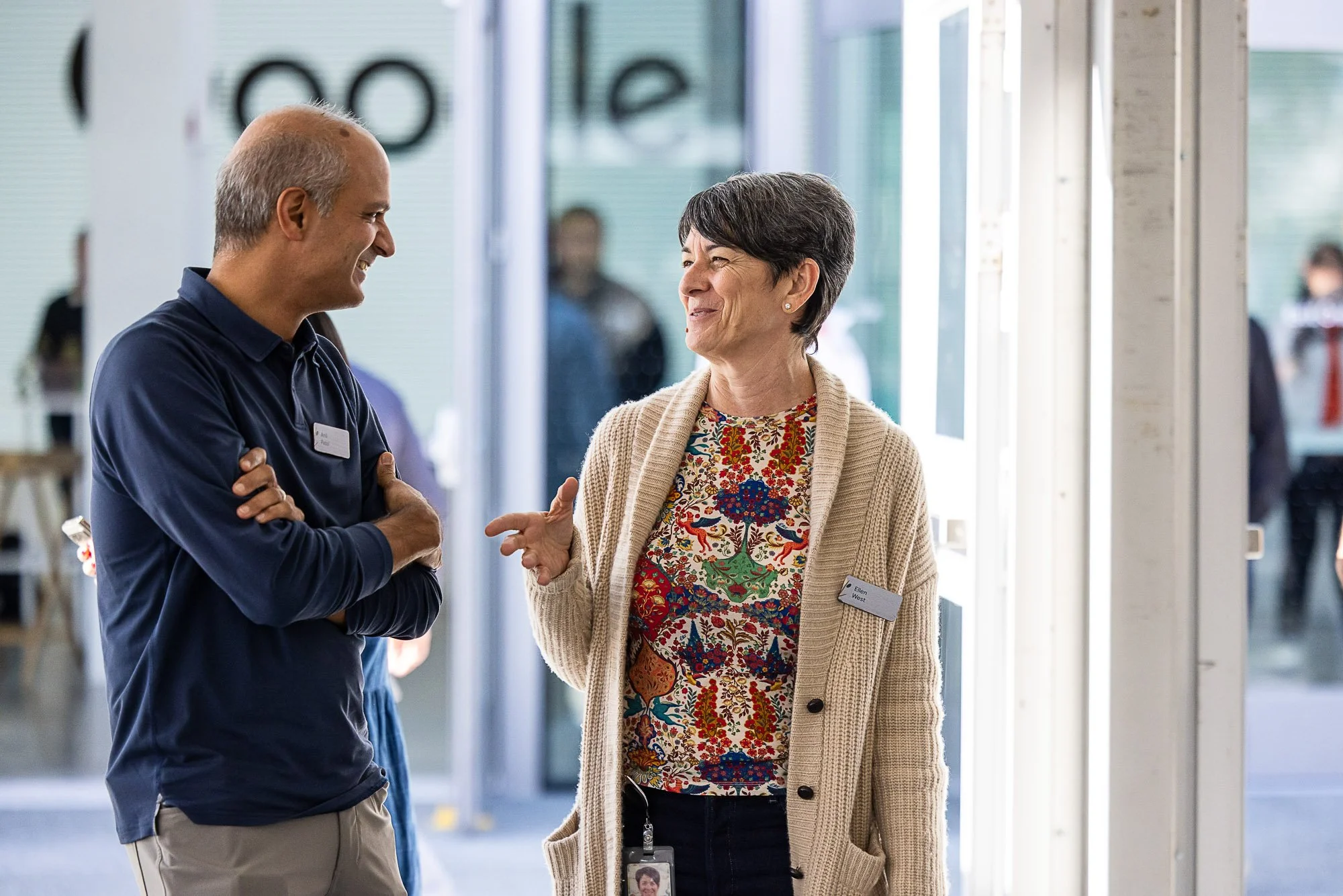 Two people, a man and a woman, are speaking to each other and smiling. The woman has short dark hair, wears a beige cardigan and a colorful top with a floral pattern, and has a name tag that says 'Ellen West'. The man has a bald head, is dressed in a