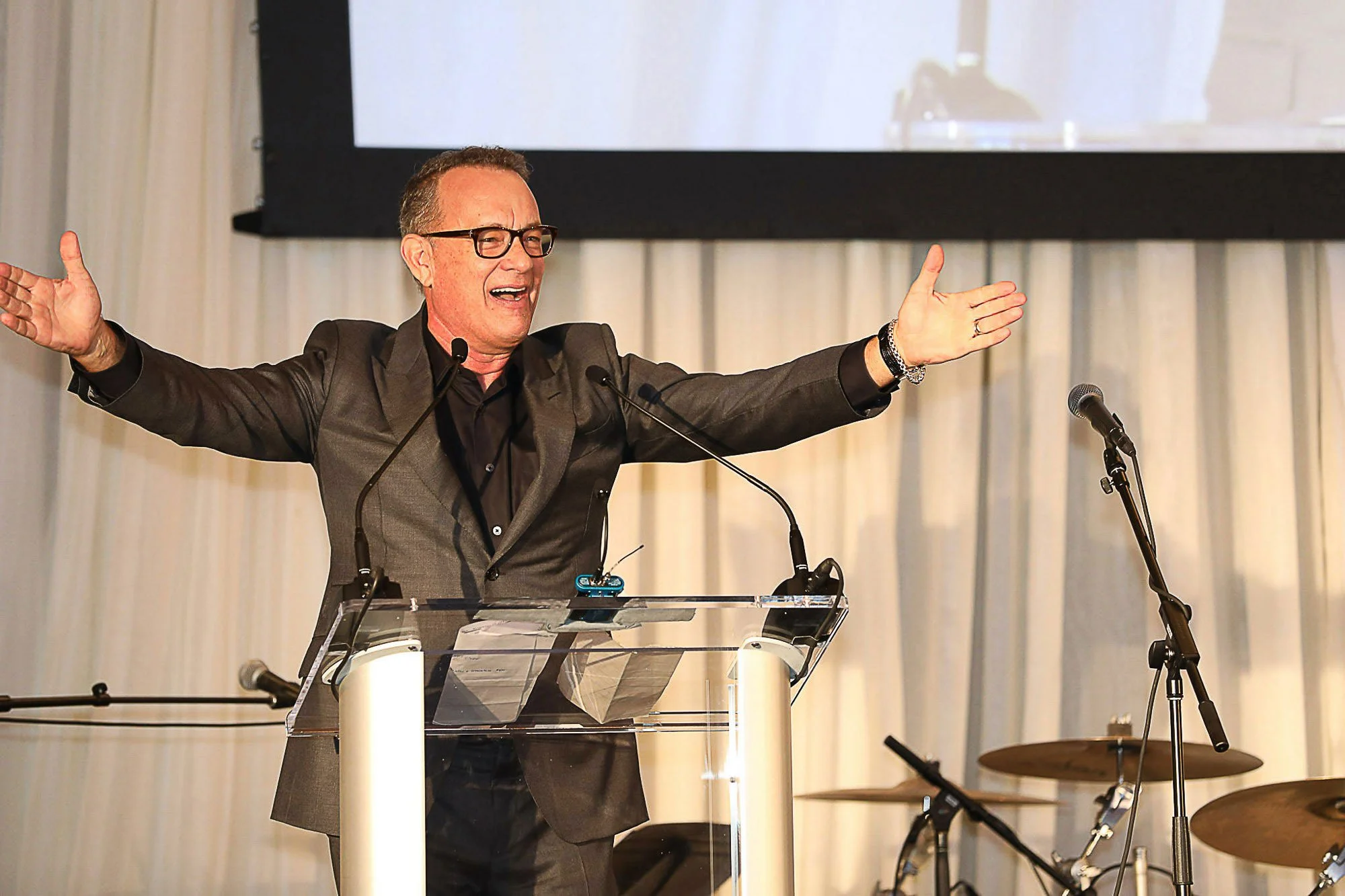 Tom Hanks in a gray suit and glasses speaking at a podium with his arms outstretched, in front of a microphone, with musical instruments visible on the stage.