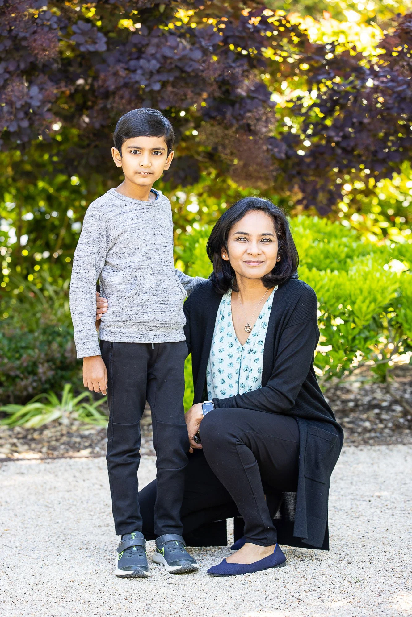 A woman with dark hair kneeling on one knee outdoors, smiling, with a young boy standing beside her. They are in a garden with green and purple foliage in the background, on a sunny day.